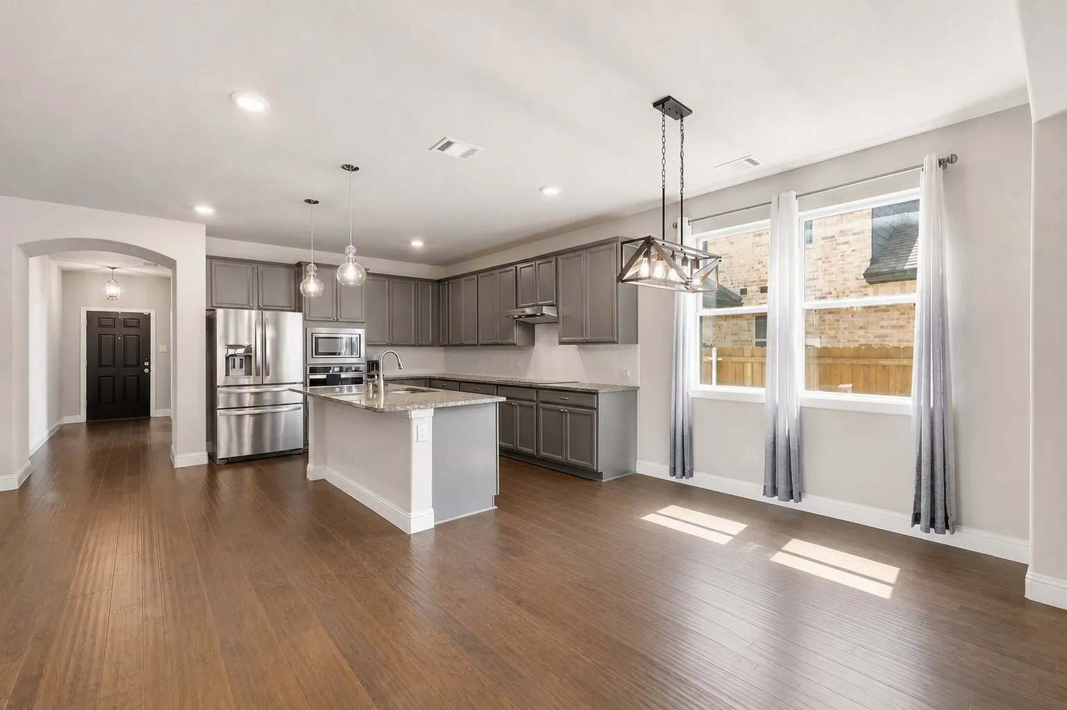 Spacious kitchen and dining area featuring wood-finish flooring, grey cabinetry, stainless steel appliances, a kitchen island with seating, and ample natural light from multiple windows