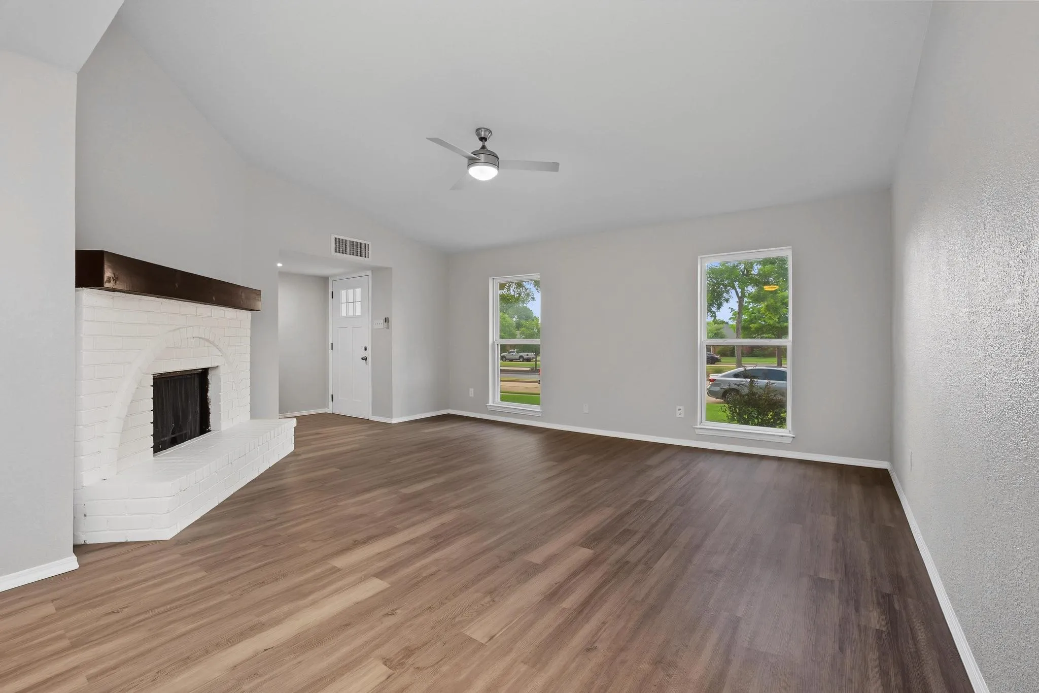 Spacious living area featuring high vaulted ceilings, wood-finish flooring, a white brick fireplace with a dark wood mantel, a ceiling fan with integrated lighting, and multiple windows