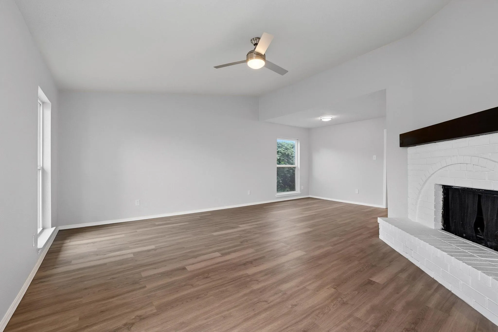Living area featuring wood-finish flooring, a white brick fireplace with a dark wood mantel, and a ceiling fan with integrated lighting