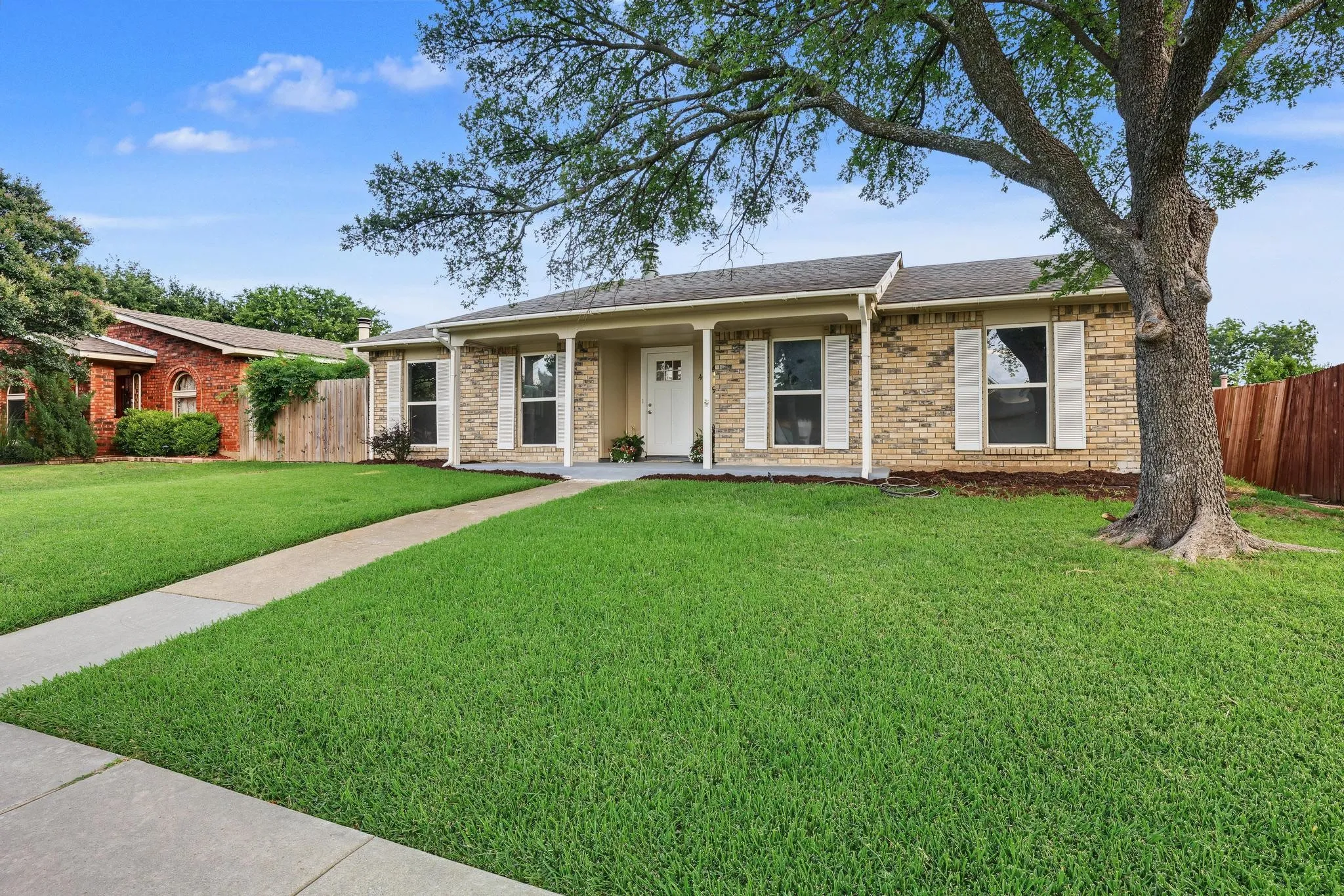 Brick exterior home featuring a covered front porch, white window shutters, and a prominent mature tree in the front yard