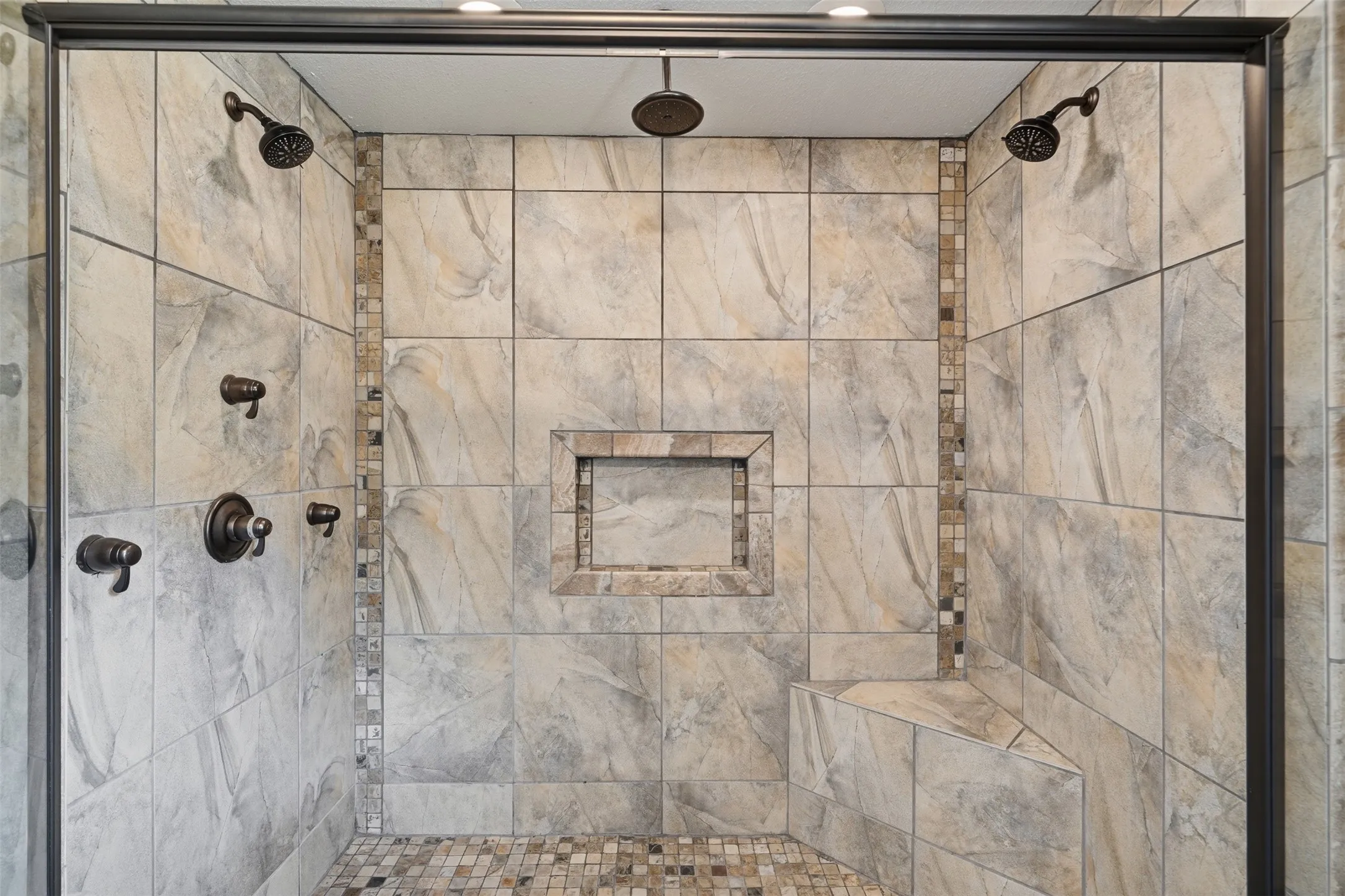 Tiled shower featuring multiple showerheads, a built-in bench, a recessed niche, and mosaic accent tiling
