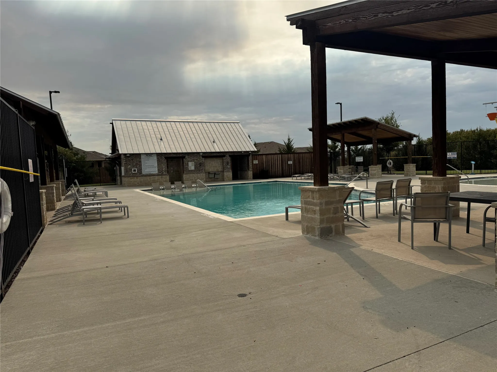 Community swimming pool with a stone-accented building featuring a metal roof, an adjacent covered pergola with stone columns, and a secondary covered structure