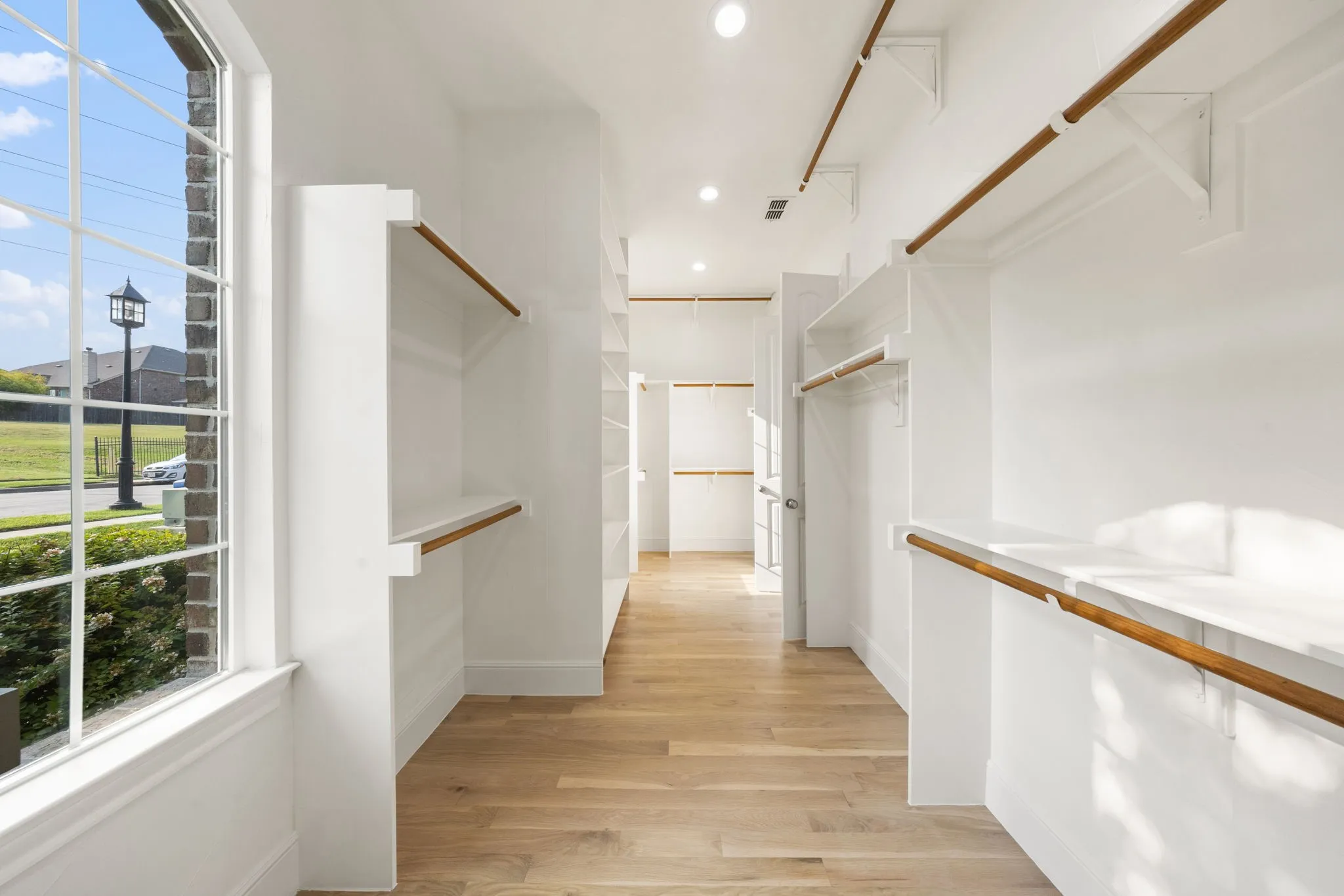 Spacious walk-in closet featuring light wood-finish flooring, recessed lighting, built-in shelving, hanging rods, and a large multi-pane window