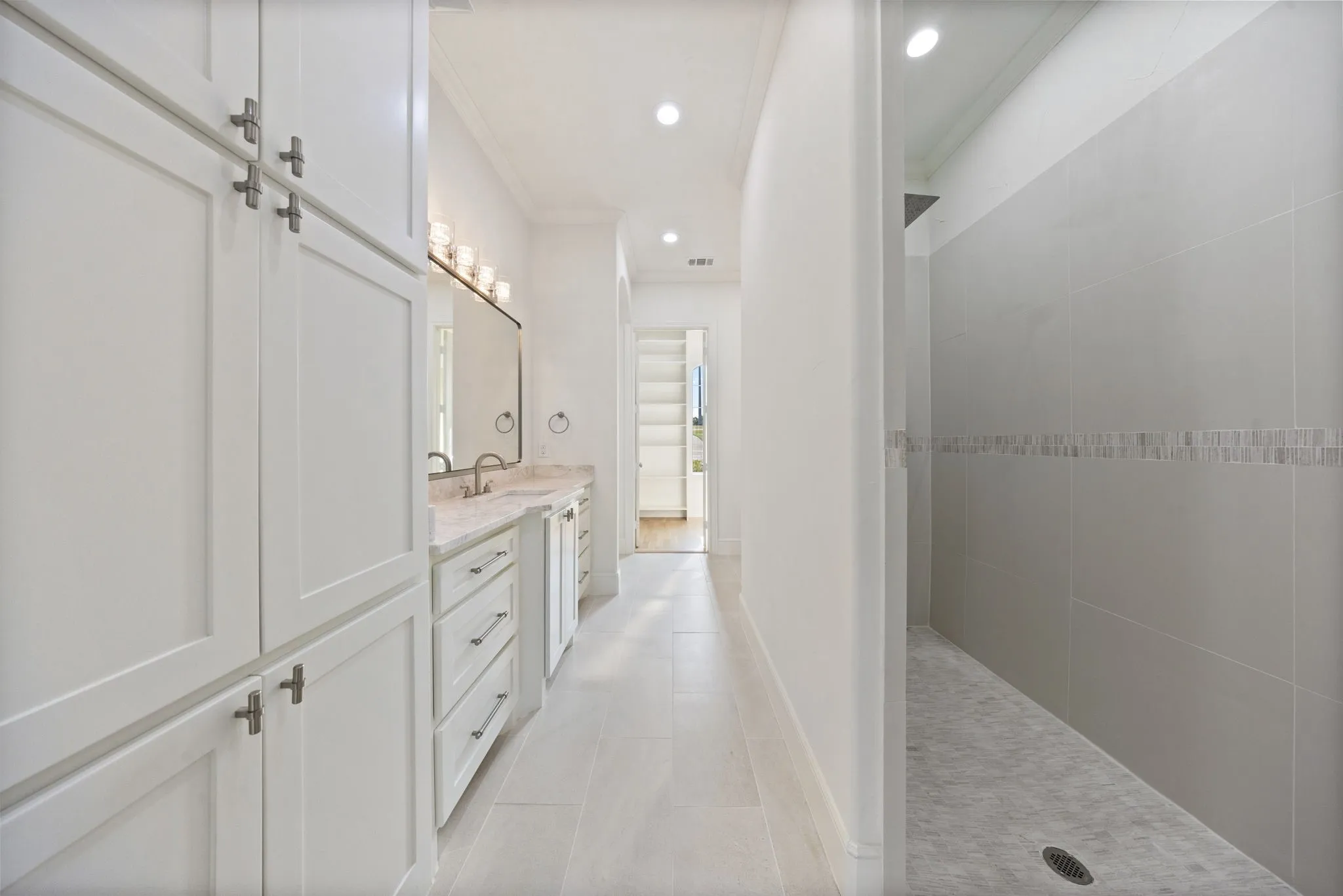 Spacious bathroom featuring white shaker cabinetry, an extended dual vanity with a light-toned countertop, and a walk-in shower with tiled walls and a decorative accent band