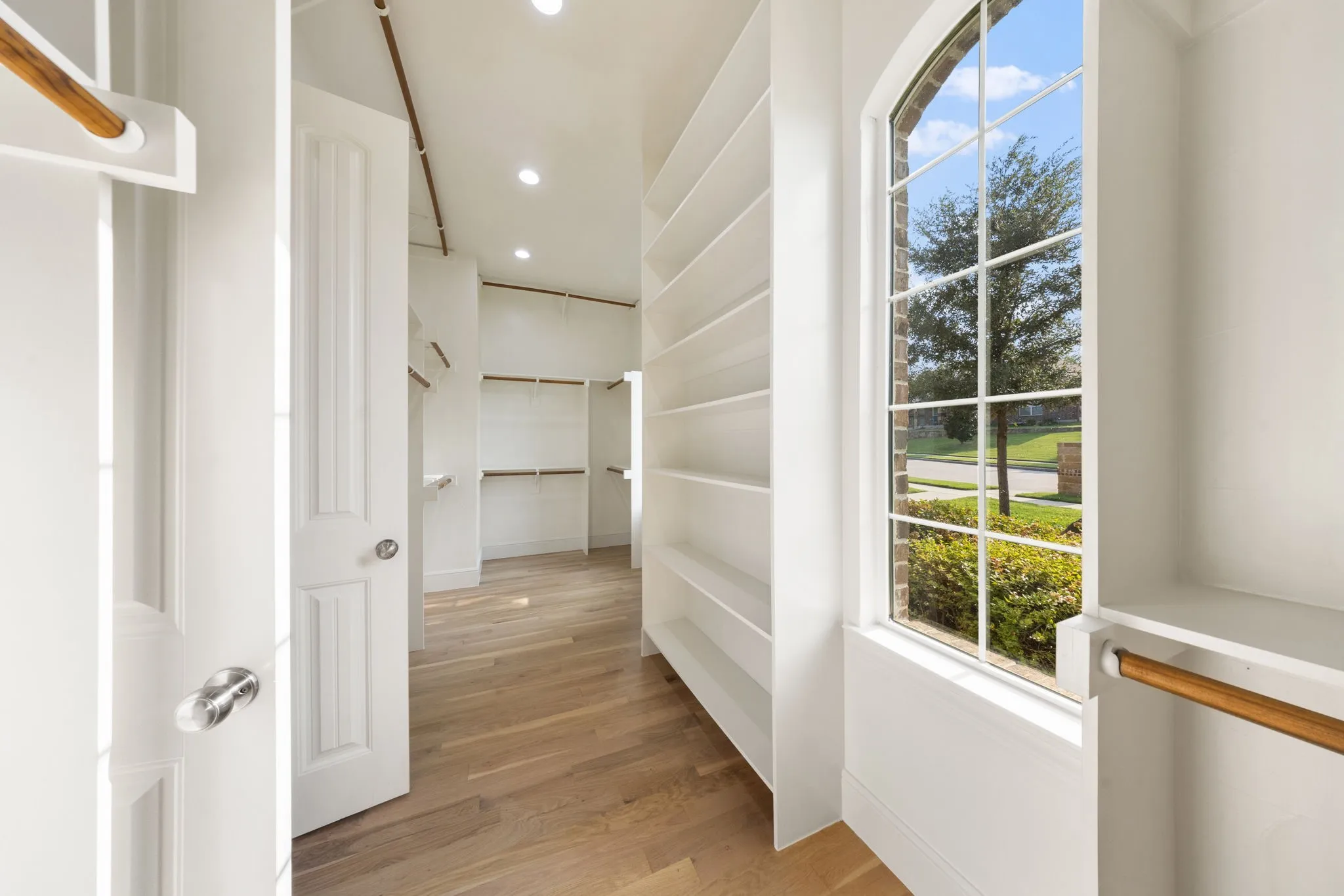 Spacious walk-in closet featuring light wood-finish flooring, recessed lighting, extensive built-in shelving, multiple hanging rods, and an arched window
