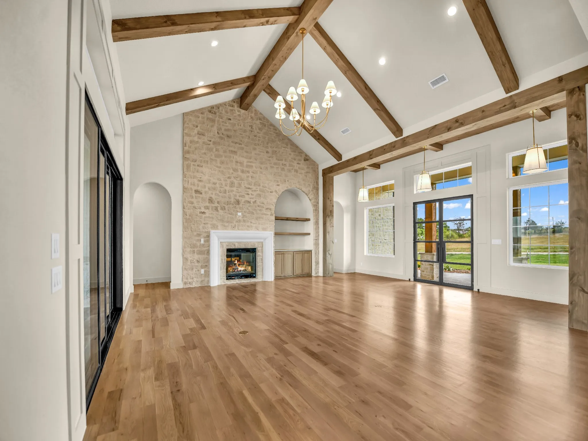 Vaulted ceiling with exposed wood beams, stone fireplace feature with mantel, wood-finish flooring, built-in shelving, and multiple large windows