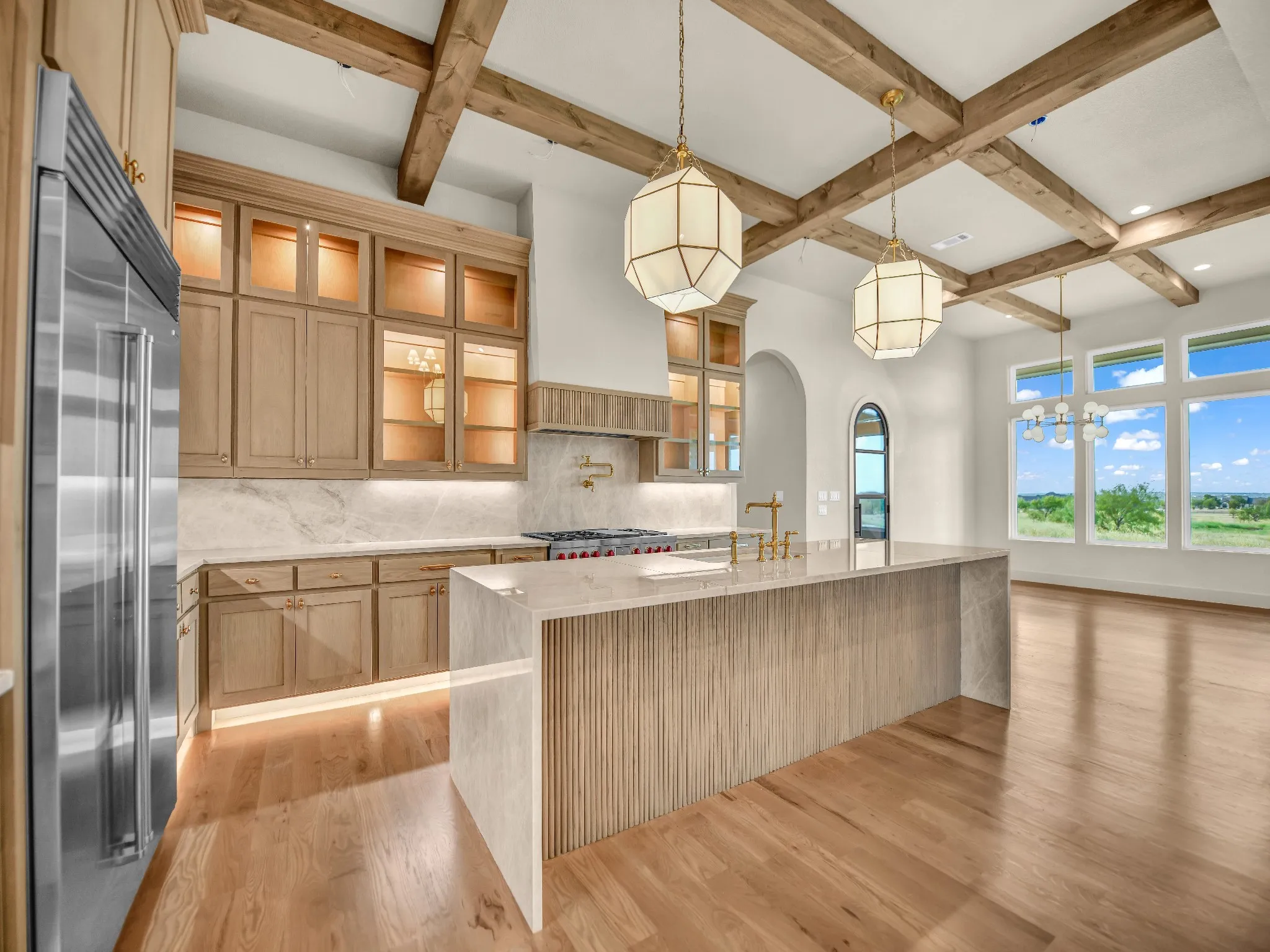 Kitchen featuring exposed wood beams, light wood-finish cabinetry with glass accents, a large center island with fluted detailing, and stainless steel appliances