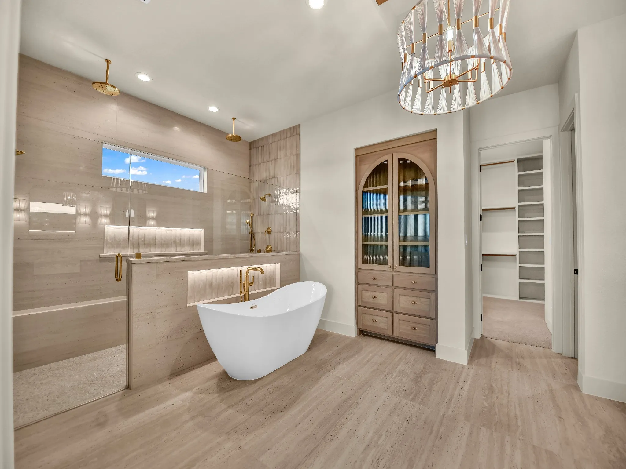 Freestanding soaking tub, walk-in shower with dual rain showerheads and gold-finish fixtures, tile flooring, built-in cabinetry, and a contemporary chandelier