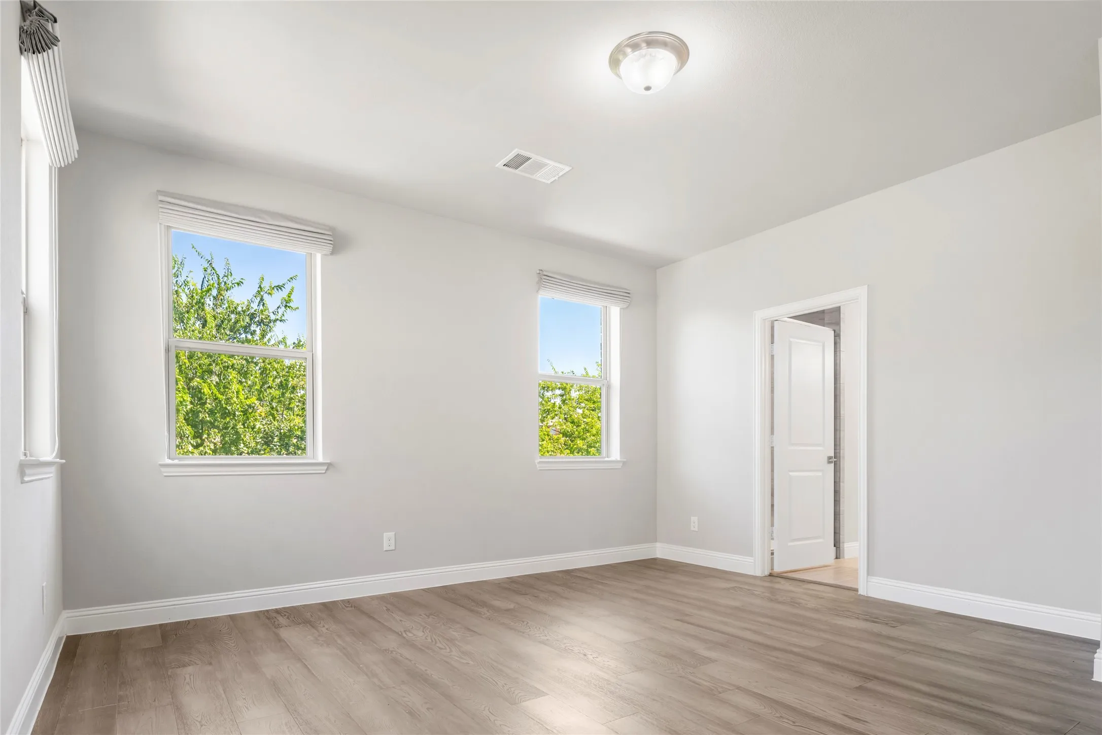 Spacious room featuring two large windows, wood-finish flooring, light gray walls, white trim, and a ceiling-mounted fixture