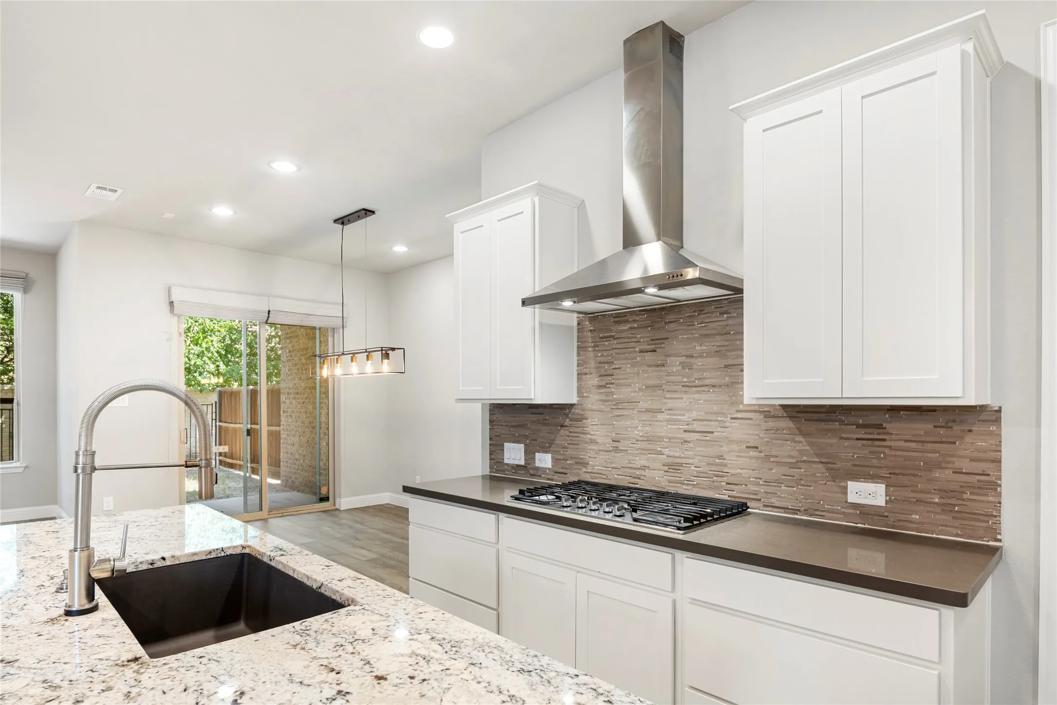 Modern kitchen featuring a stainless steel range hood, stone tile backsplash, white shaker cabinetry, and light-toned granite countertops