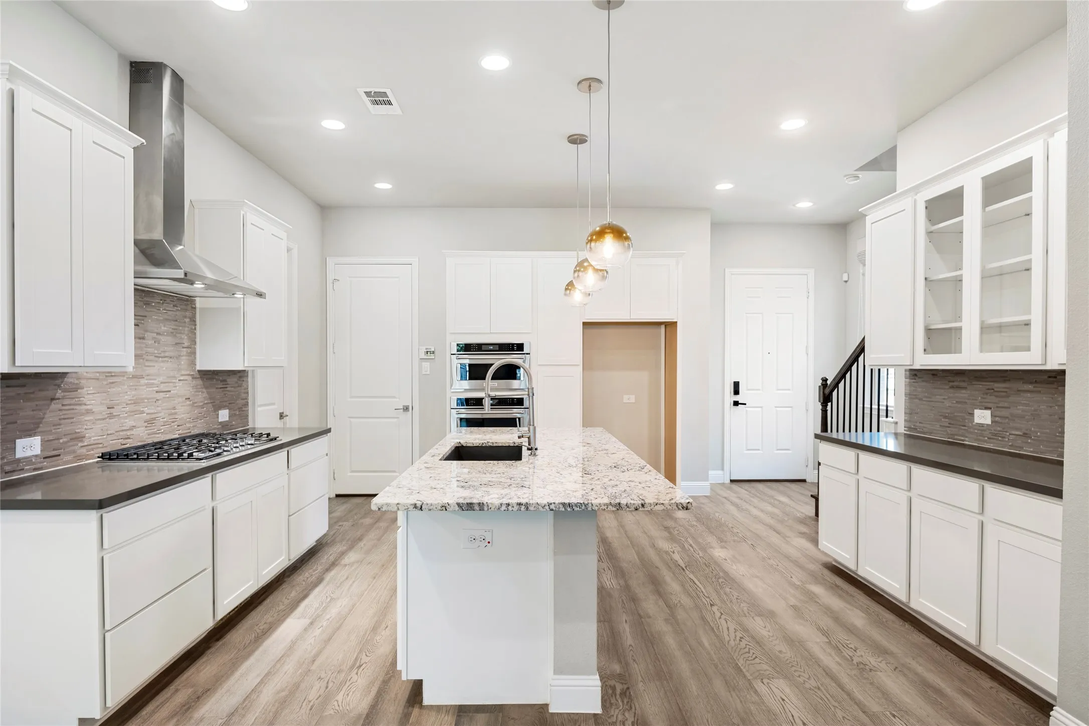 Kitchen featuring a central island with a light-toned countertop and pendant lighting, white cabinetry, wood-finish flooring, a stainless steel range hood, and a built-in oven