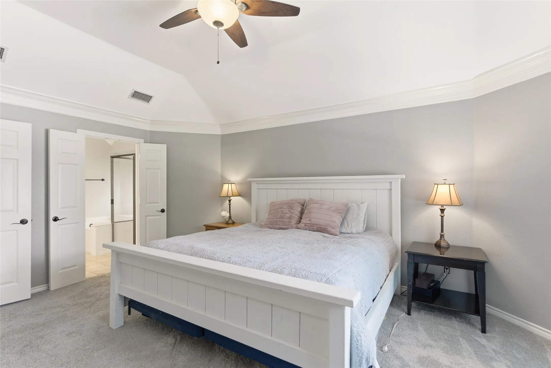 Vaulted ceiling with crown molding, ceiling fan, carpeted flooring, and double doors opening to a bathroom featuring a built-in tub and shower enclosure