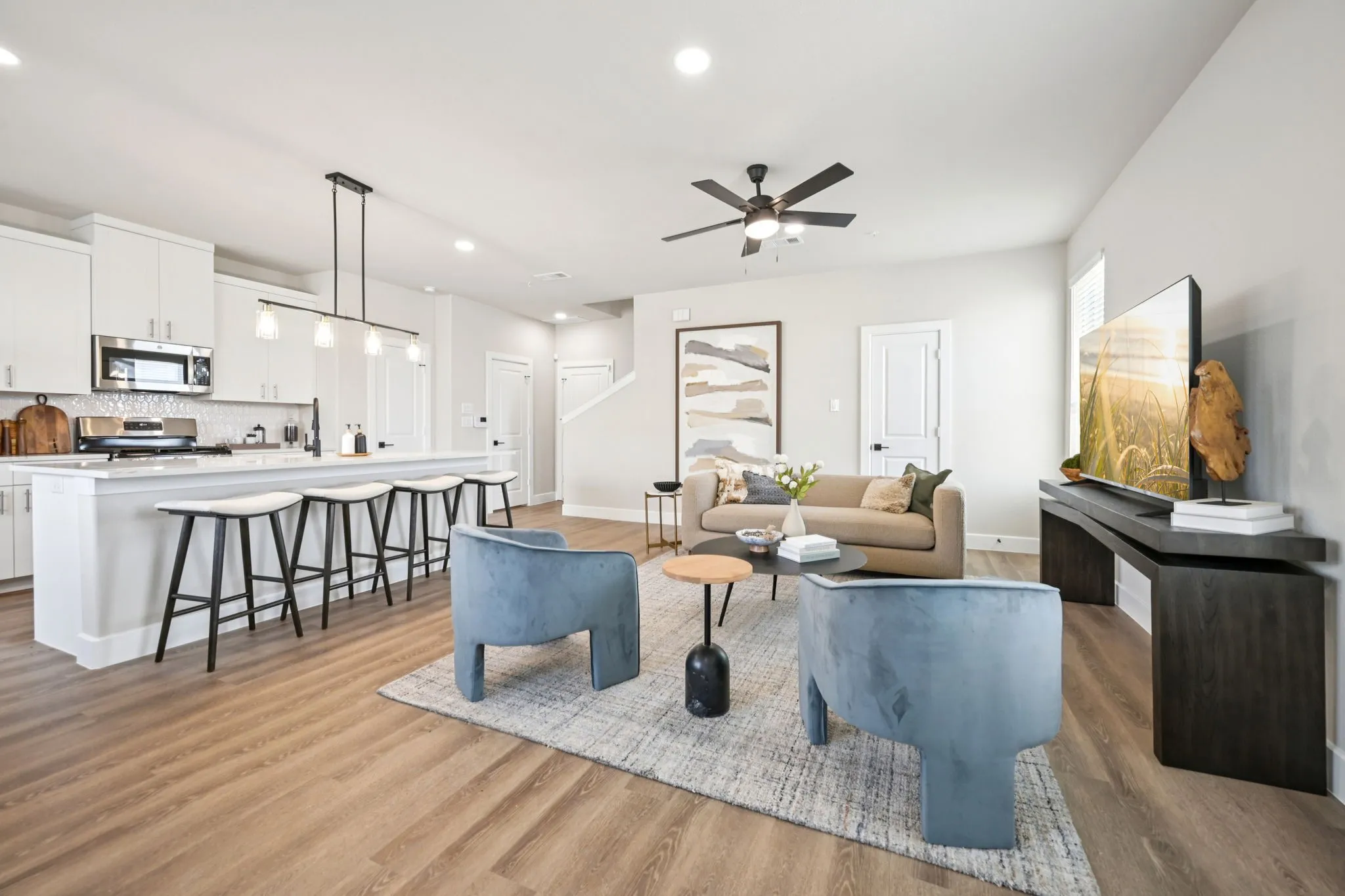 Open-concept living area featuring wood-finish flooring, a contemporary kitchen island with pendant lighting, white cabinetry, stainless steel appliances, and recessed ceiling lights