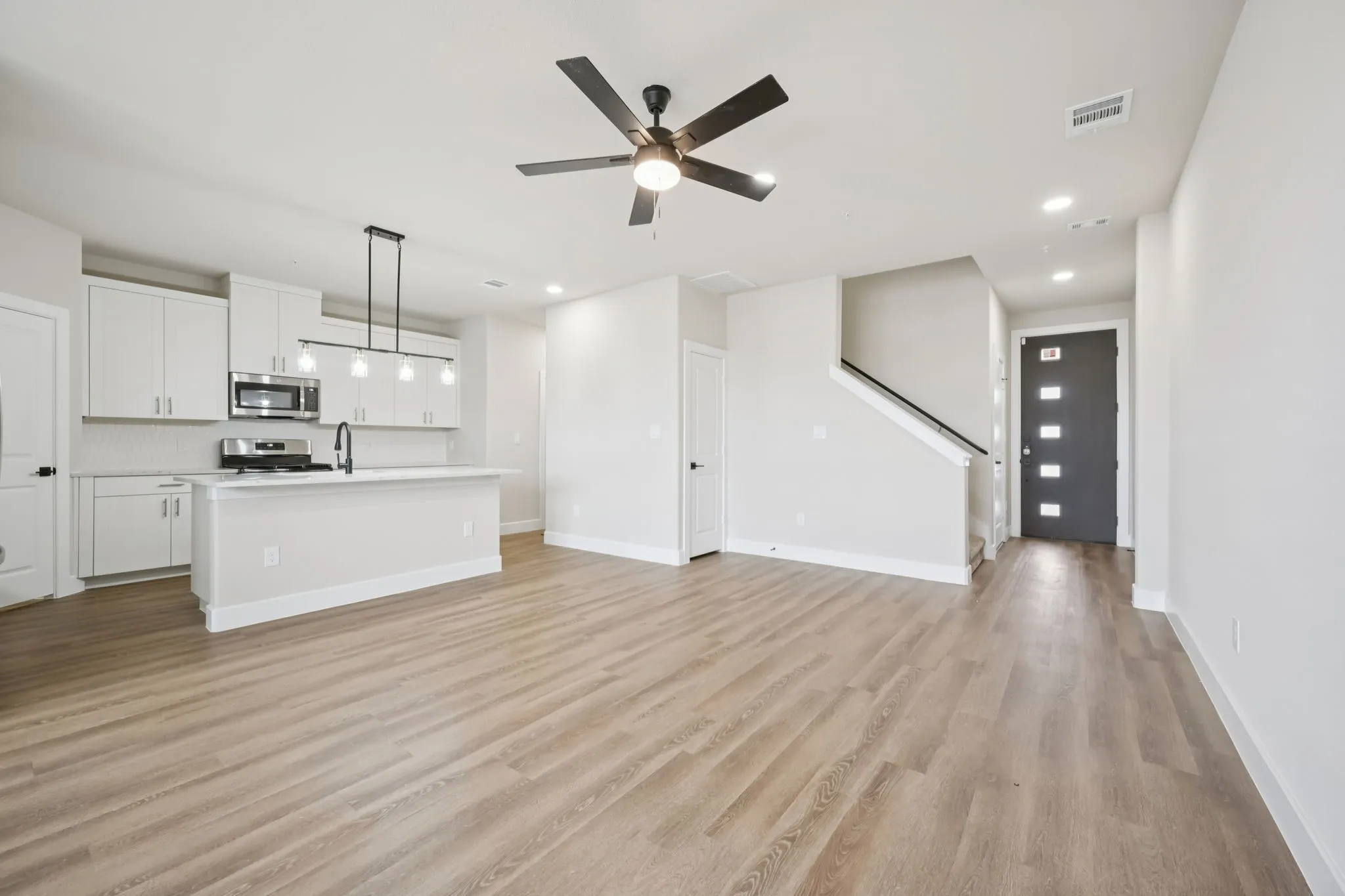 Open-concept living space featuring wood-finish flooring, a modern kitchen with white cabinetry and stainless steel appliances, a kitchen island with a sink, a contemporary linear pendant light fixture, and a ceiling fan