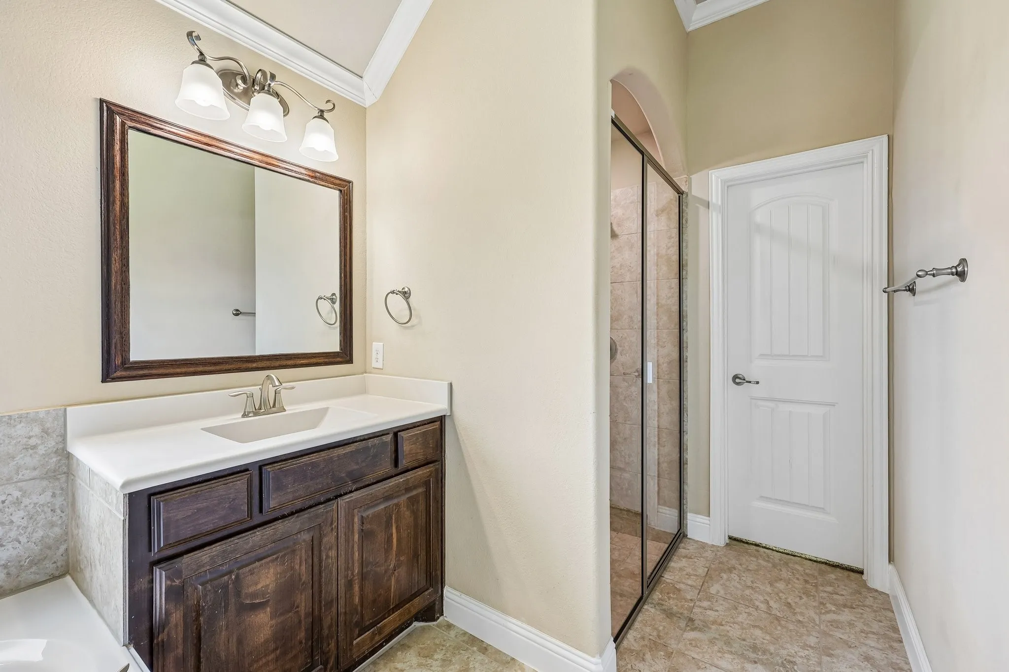 Bathroom featuring a dark wood vanity with a white countertop and undermount sink, framed mirror, three-light vanity fixture, and a glass-enclosed shower with tiled walls