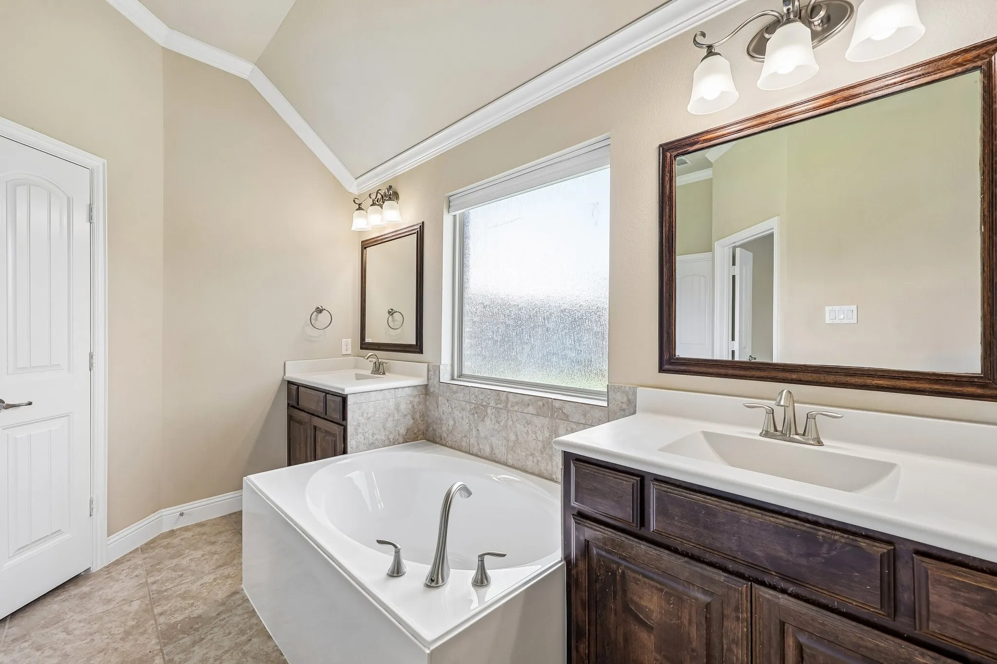 Bathroom featuring a built-in soaking tub, dual vanity sinks with white countertops, dark wood-finish cabinetry, and a textured privacy window