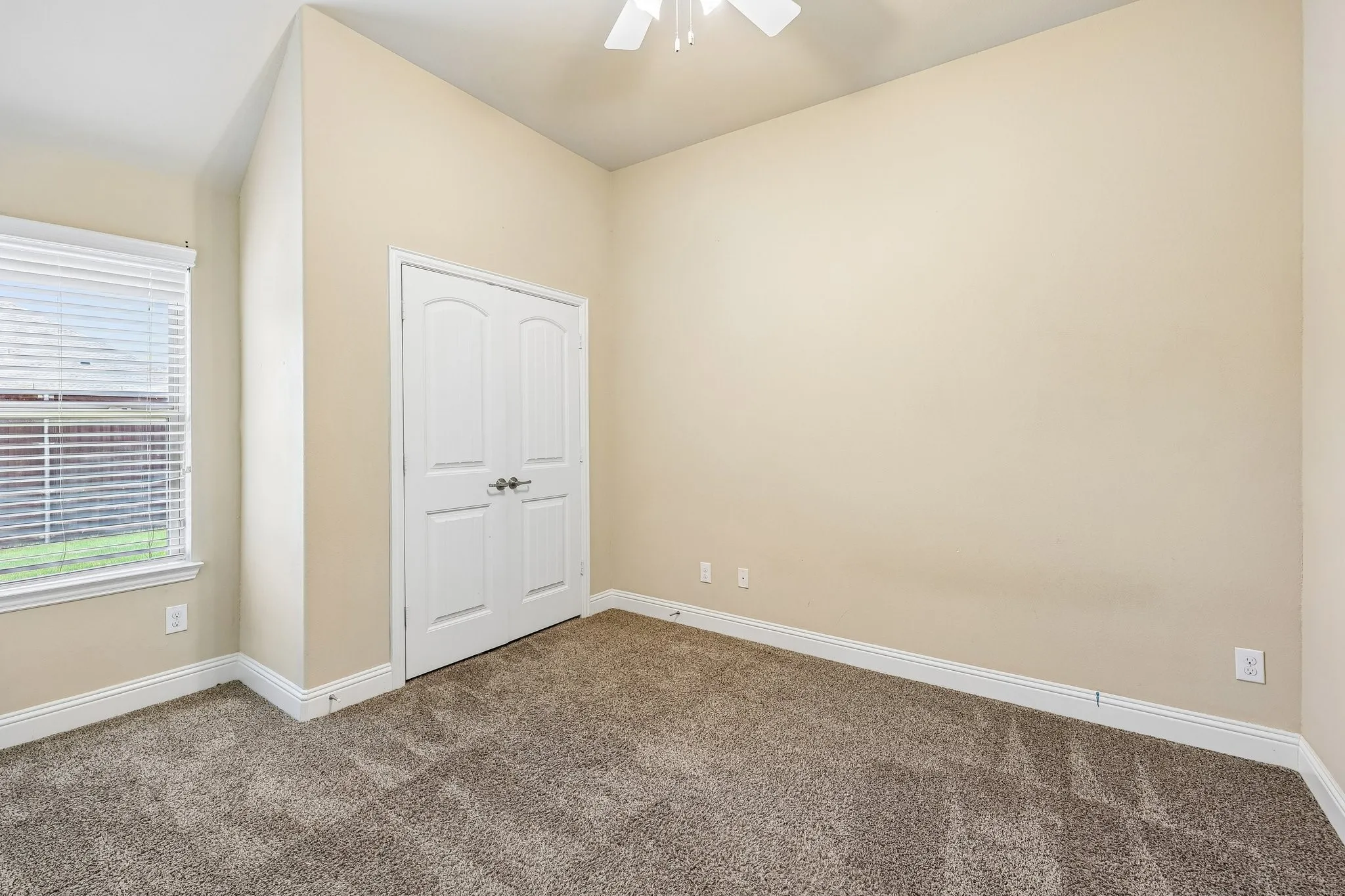 Spacious room featuring neutral-toned walls, plush carpet flooring, a double-door closet with arched paneling, a window with horizontal blinds, and a ceiling fan