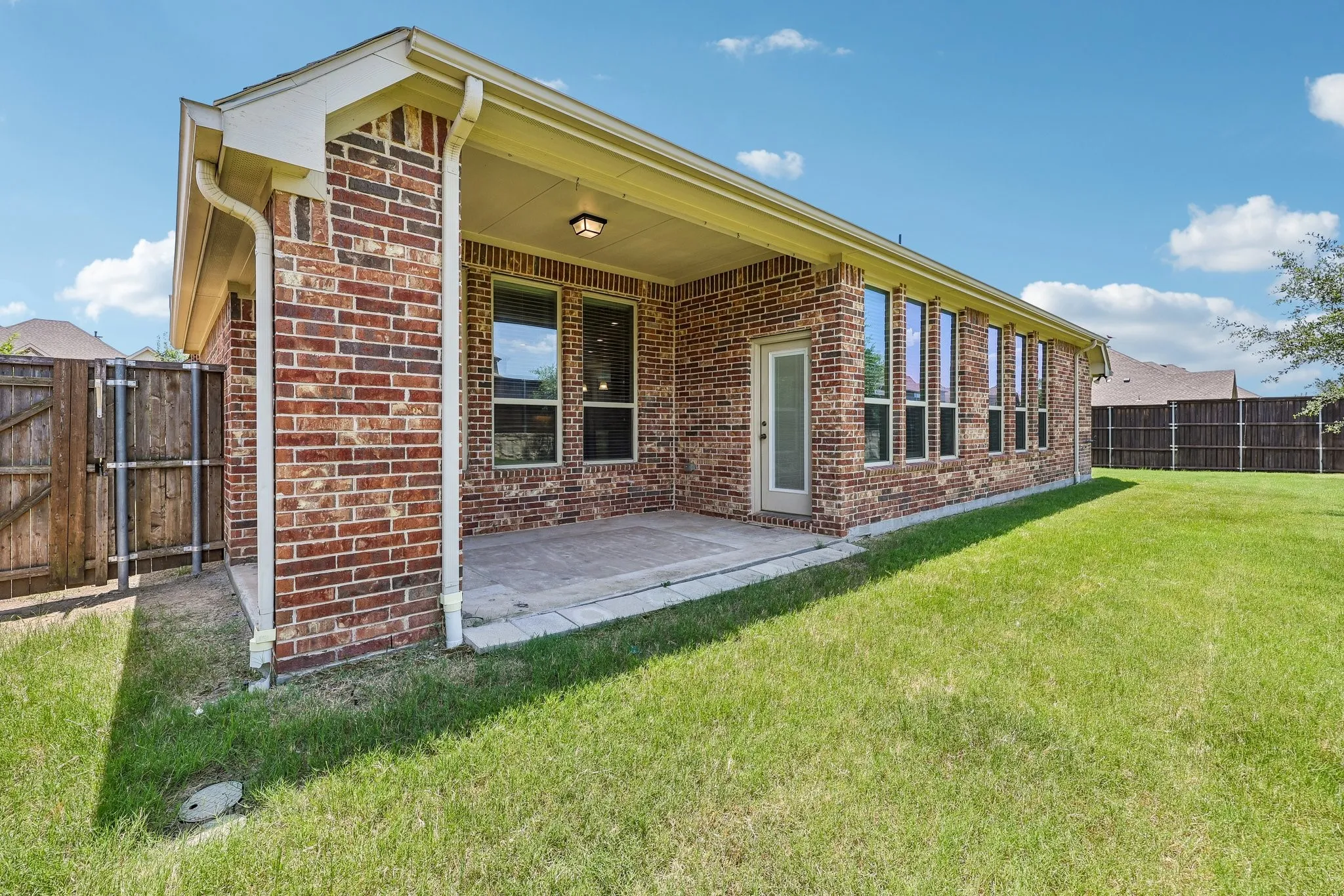 Expansive backyard featuring a covered patio, brick exterior, multiple windows, a white framed door, and a full perimeter fence