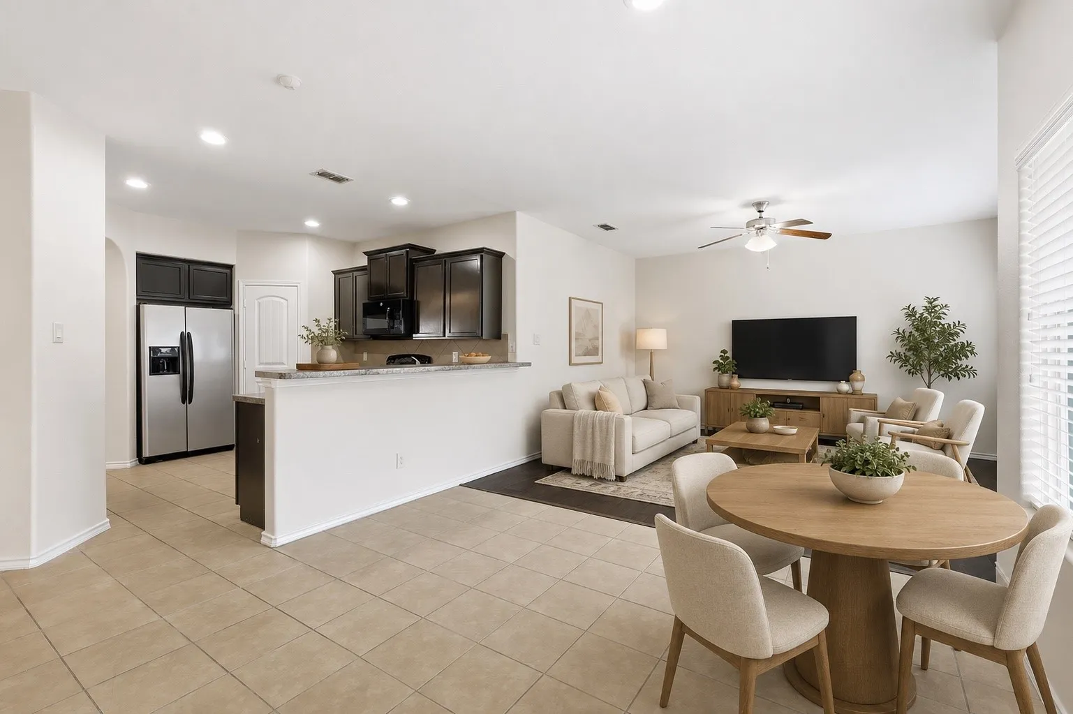 Open-concept living space featuring light-toned tile flooring, recessed lighting, a ceiling fan, a kitchen pass-through with a stone-finish countertop, and dark wood cabinetry in the kitchen area