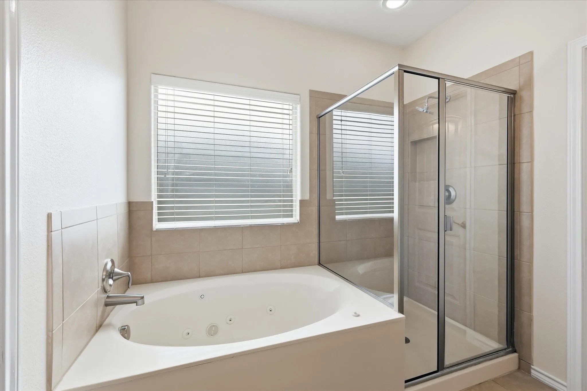 Bathroom featuring a built-in jetted tub, a glass-enclosed shower, neutral tile surround, and a window with blinds