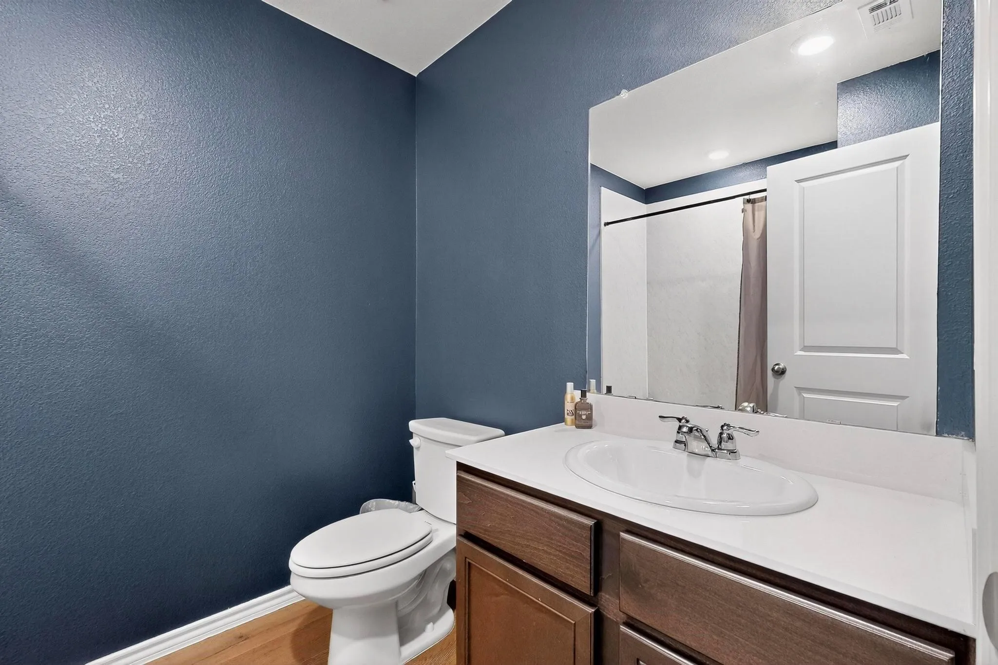 Bathroom featuring a white elongated toilet, wood-finish flooring, a vanity with a white countertop and dark wood-finish cabinetry, and a large wall mirror