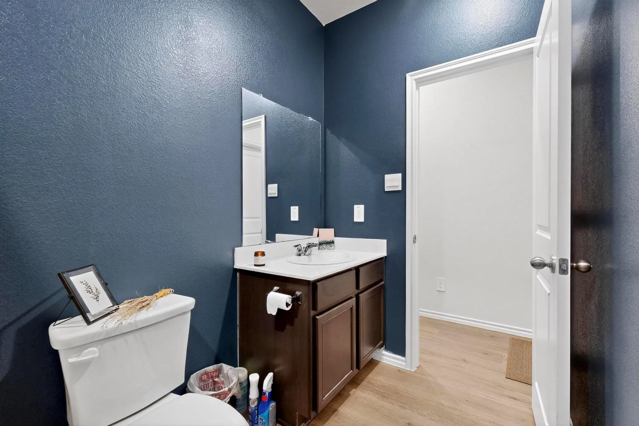 Powder room featuring a white single-basin vanity with a dark wood-finish cabinet, a wall-mounted mirror, and wood-finish flooring