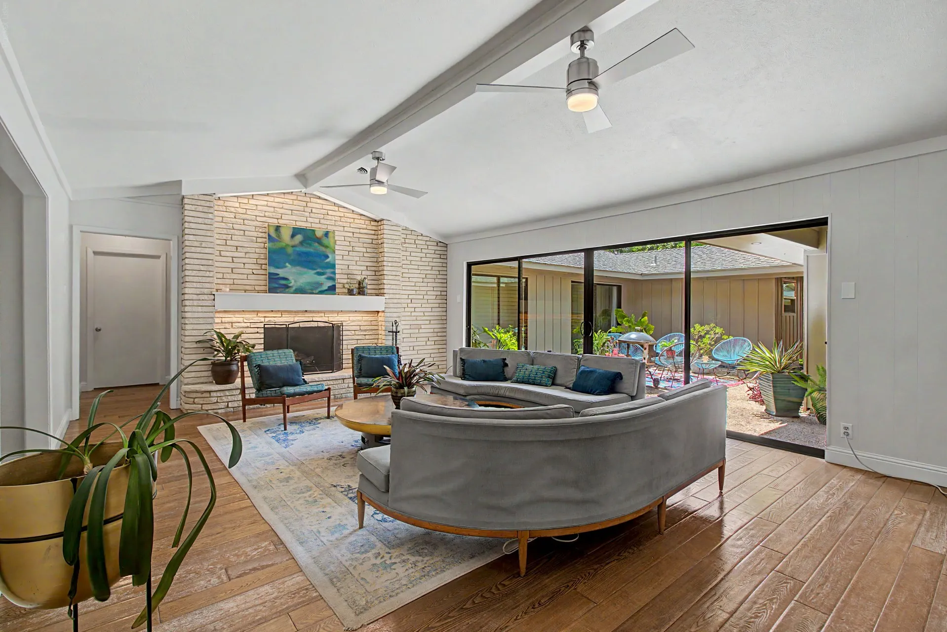Spacious living area featuring vaulted ceilings, a brick fireplace, wood-finish flooring, and large glass sliders opening to an outdoor courtyard