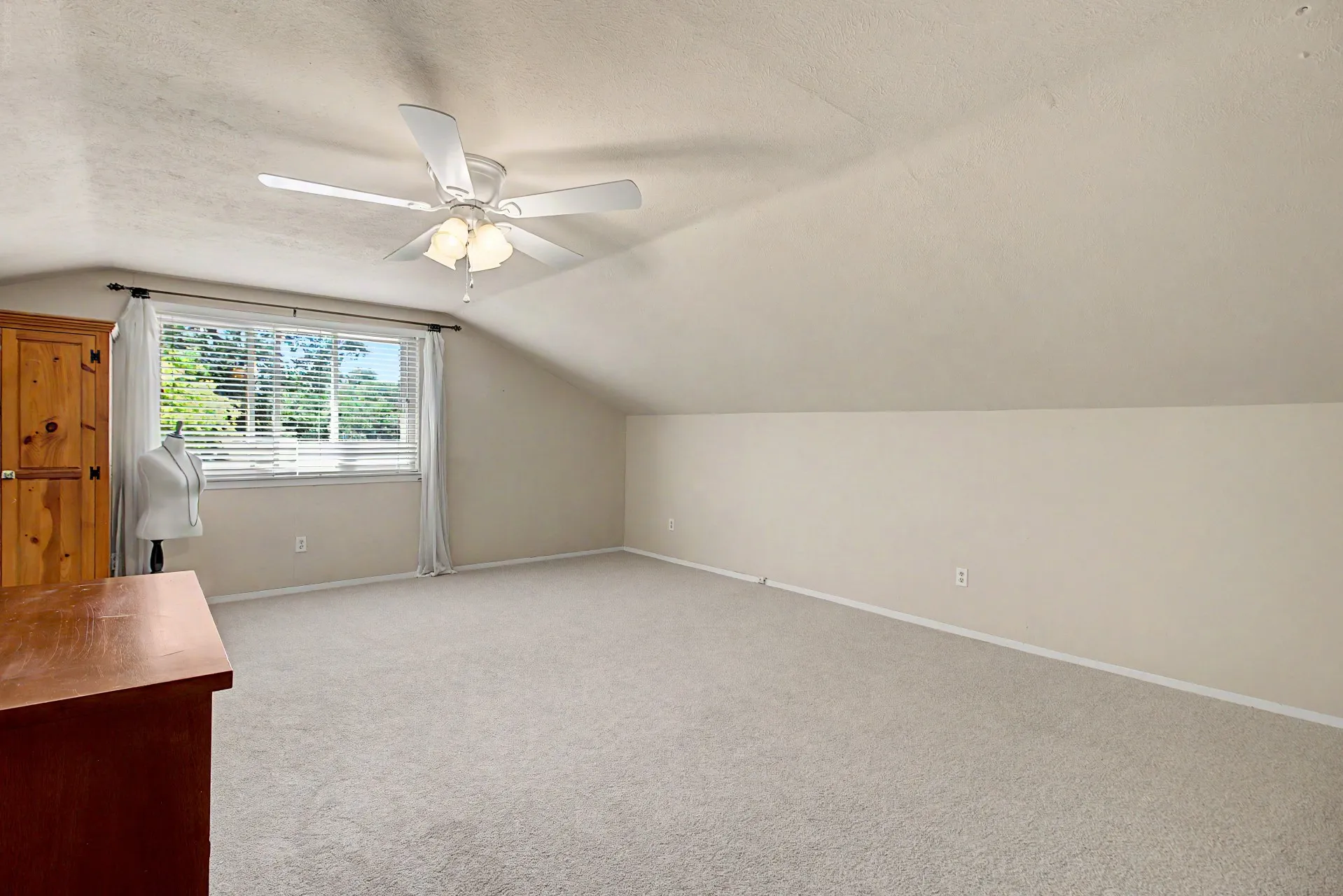 Spacious carpeted room featuring a ceiling fan with integrated lighting, a window with horizontal blinds and drapery rods, and a slanted ceiling