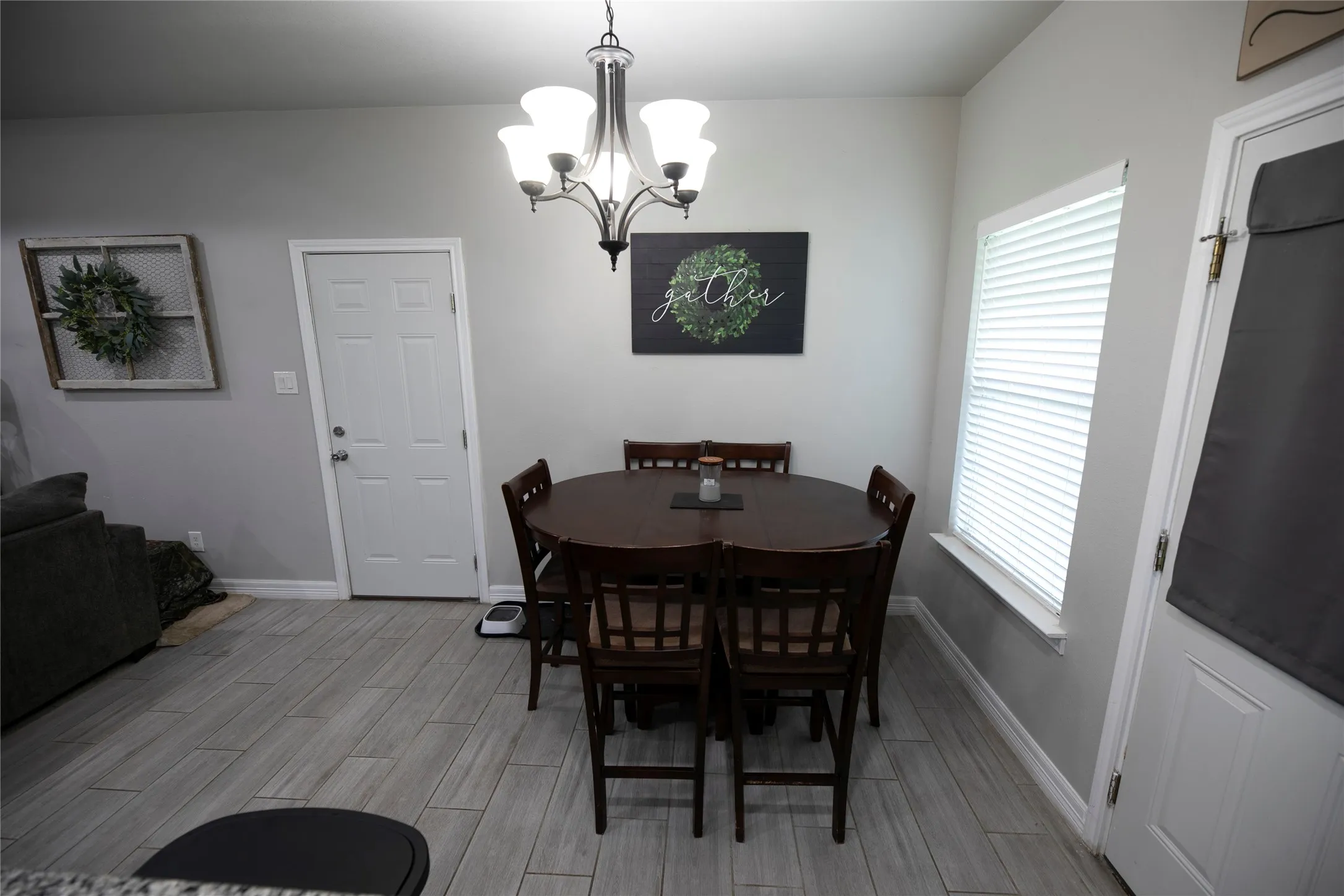 Inviting dining area featuring wood-finish tile flooring, a multi-light chandelier, a sun-filtering window with blinds, a paneled door, and neutral wall tones