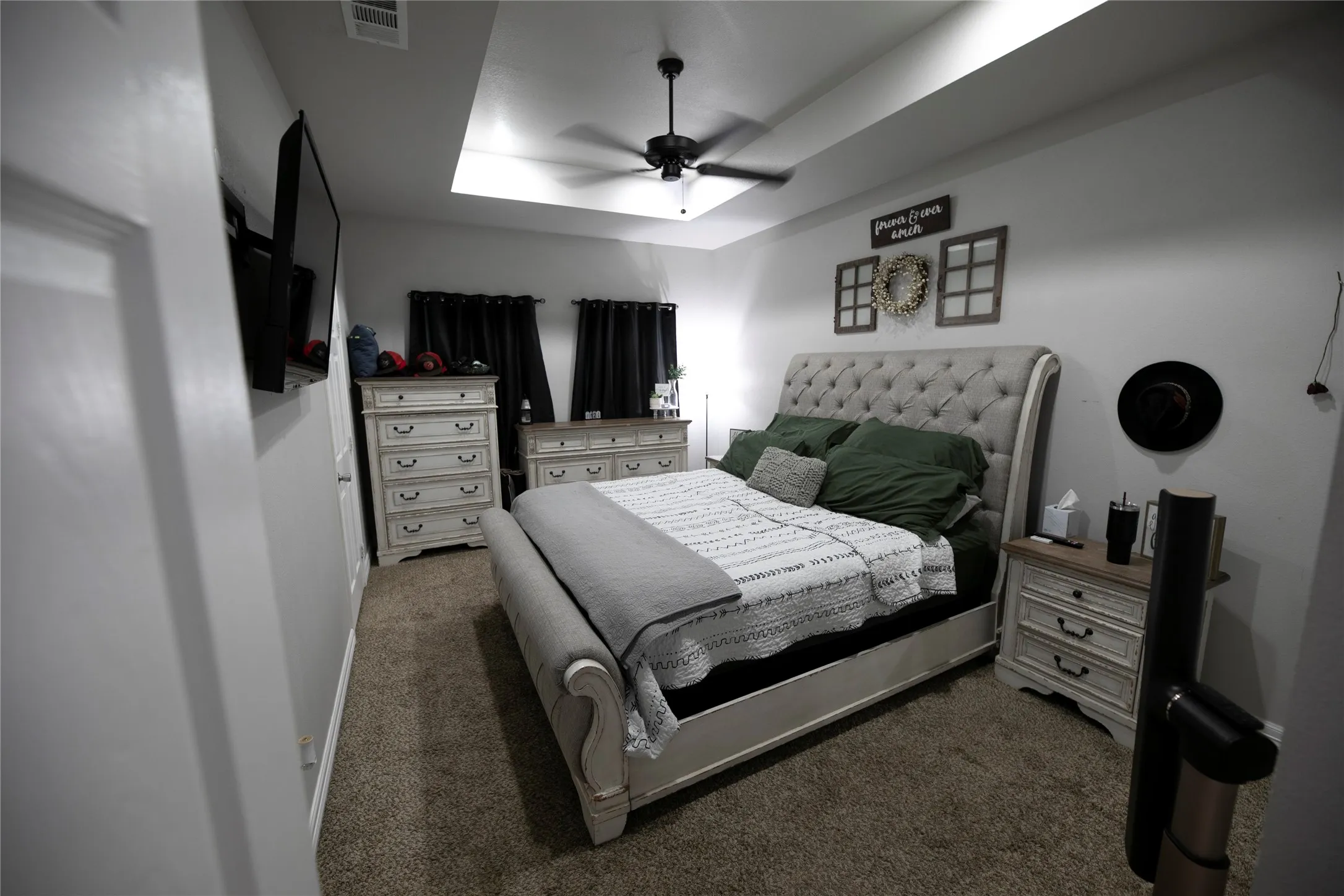 Spacious bedroom featuring a tray ceiling with a ceiling fan, neutral carpeting, and a wall-mounted television