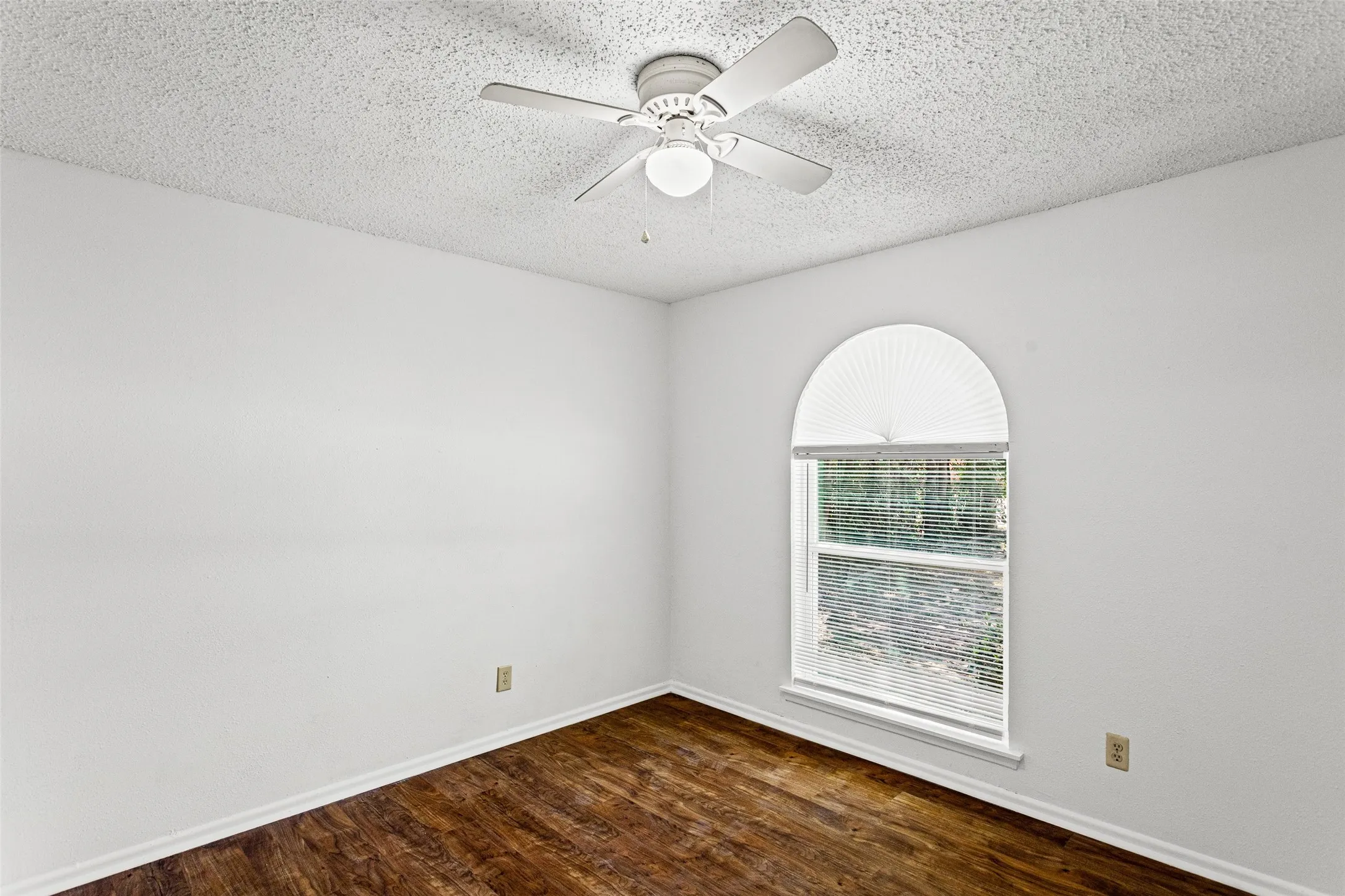 Arched window with pleated shade, wood-finish flooring, white baseboards, and a white ceiling fan with integrated lighting