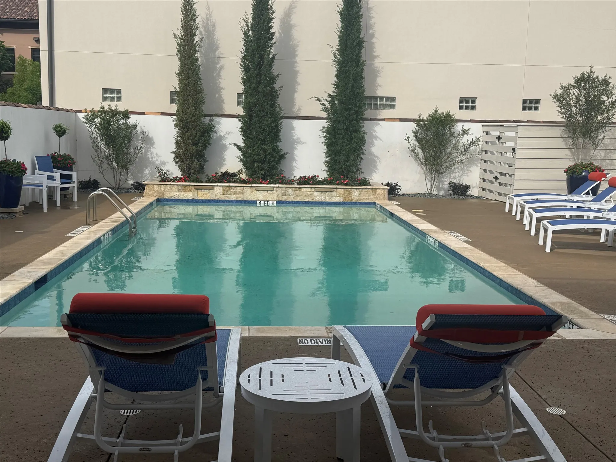 Community Pool Rectangular swimming pool featuring a light-colored coping, concrete decking, a raised planter with mature trees, and a decorative privacy screen
