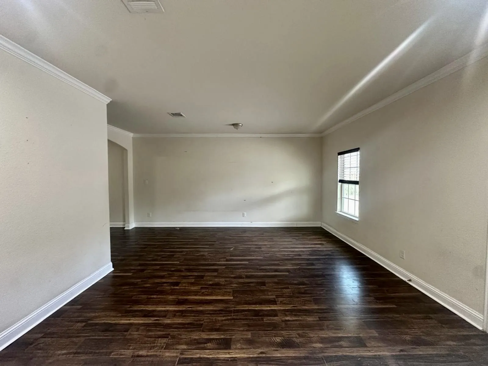 Spacious room featuring rich wood-finish flooring, crisp white baseboards, crown molding, and a window with blinds