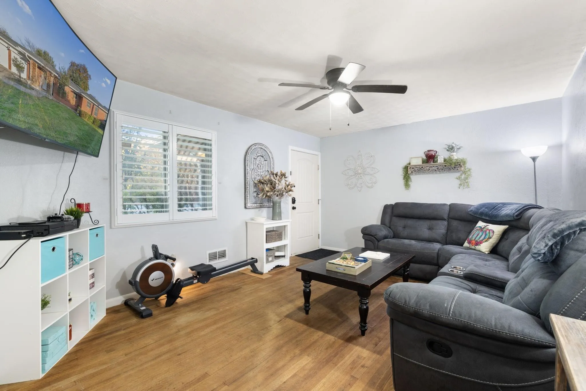Living area featuring wood-finish flooring, a ceiling fan with integrated lighting, and multiple window openings with white louvered shutters