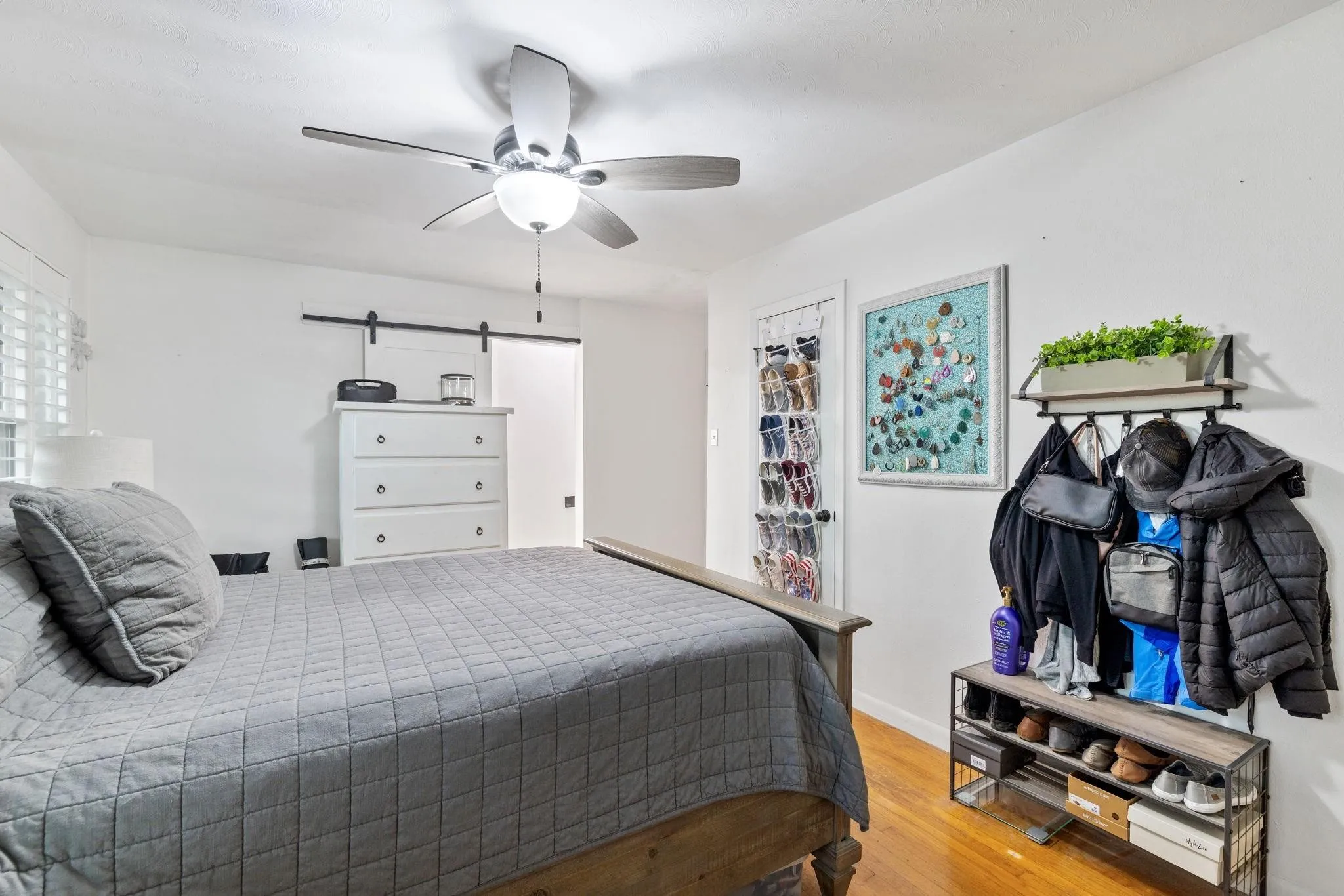 Room featuring wood-finish flooring, a ceiling fan with integrated lighting, window with white louvered shutters, and a modern barn door