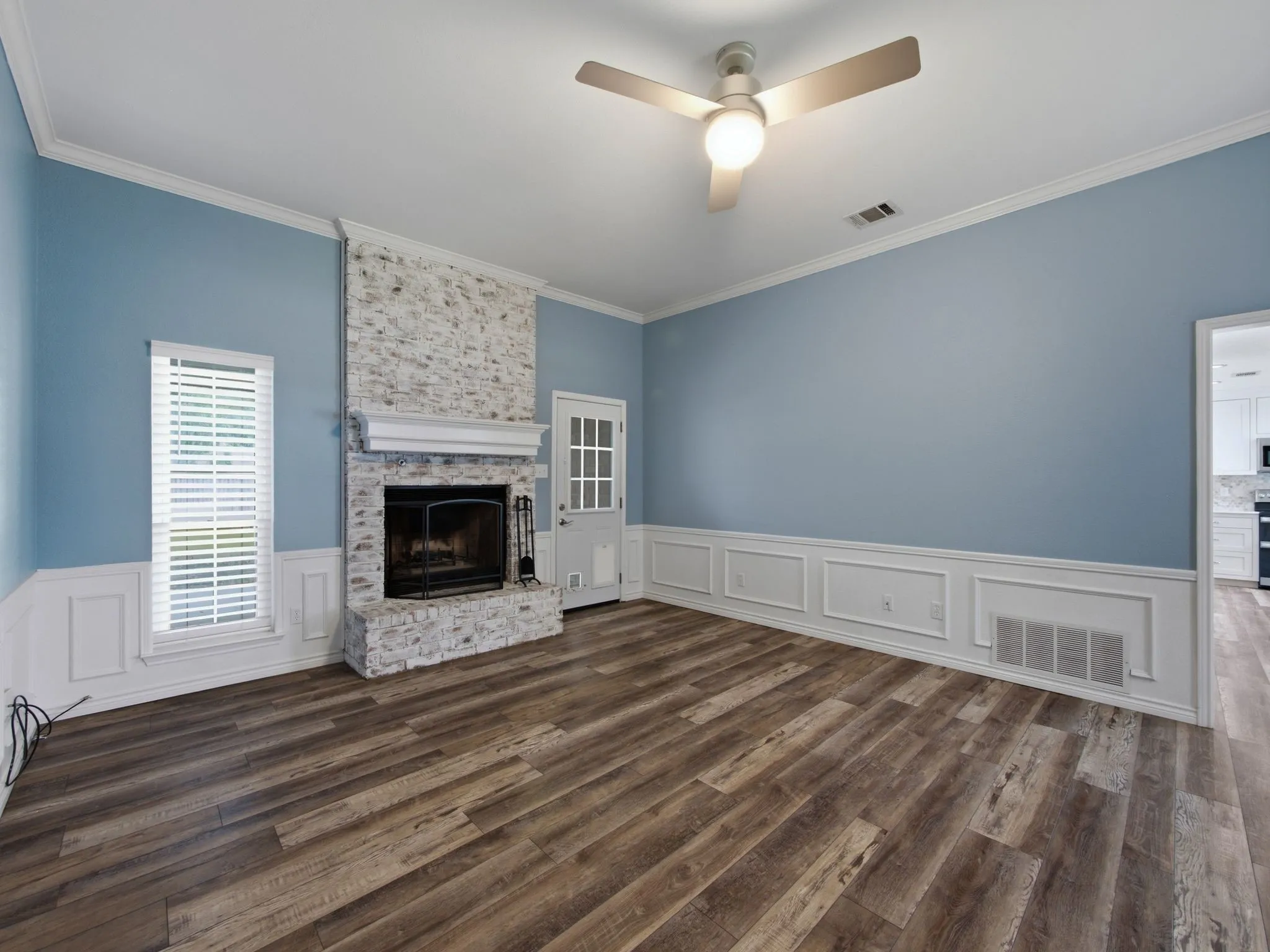 Living area featuring a white-washed brick fireplace with a mantle, wood-finish flooring, a ceiling fan and wainscoting with decorative trim