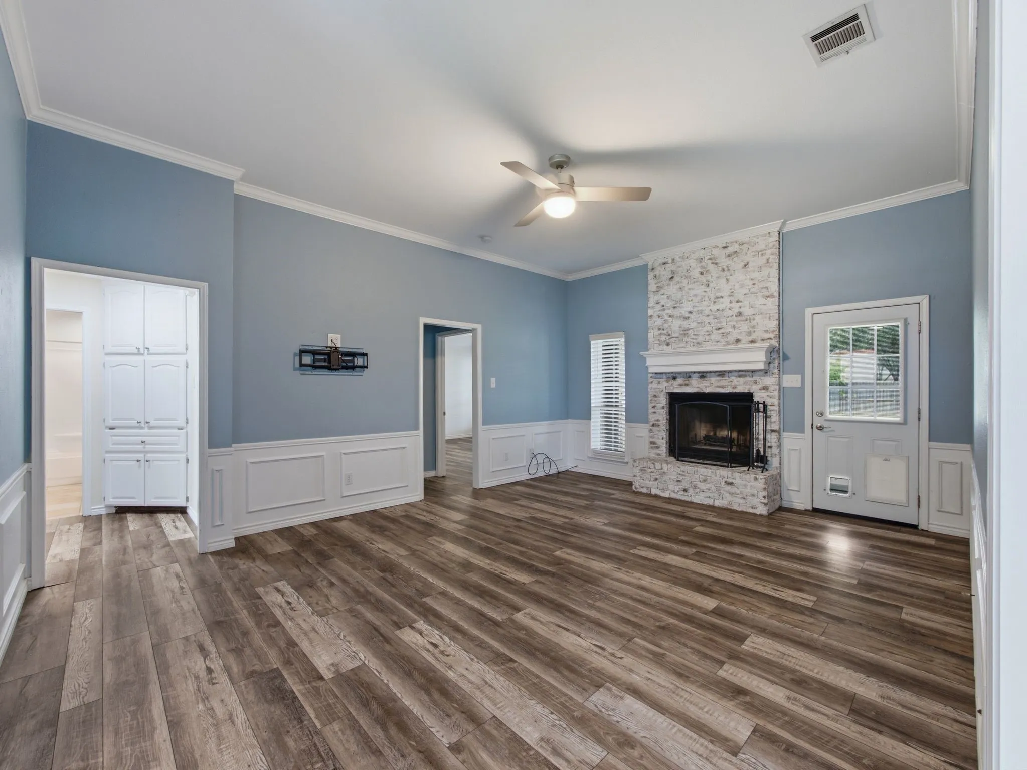 Living area featuring a white-washed brick fireplace with a mantle, wood-finish flooring, a ceiling fan and wainscoting with decorative trim