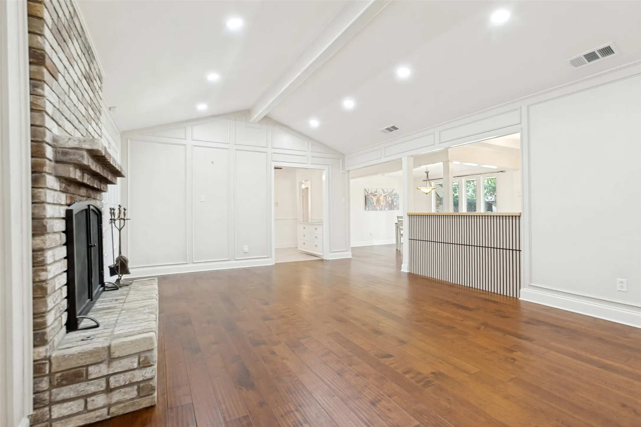 Spacious living area featuring wood-finish flooring, a brick fireplace with a stone hearth, vaulted ceilings with exposed beams, recessed lighting, and decorative wall paneling