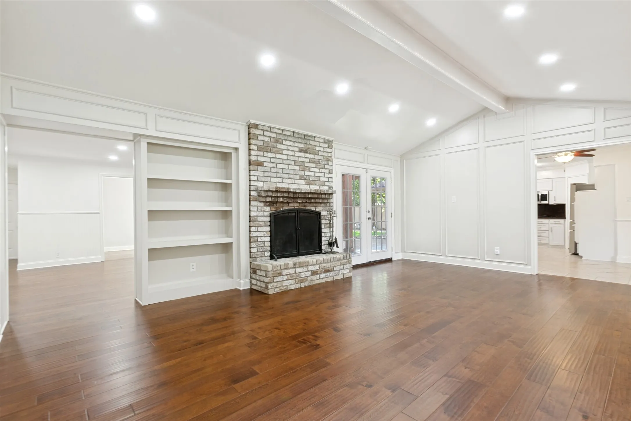 Living space featuring wood-finish flooring, a stone fireplace, built-in shelving, white paneling, and a vaulted ceiling with recessed lighting