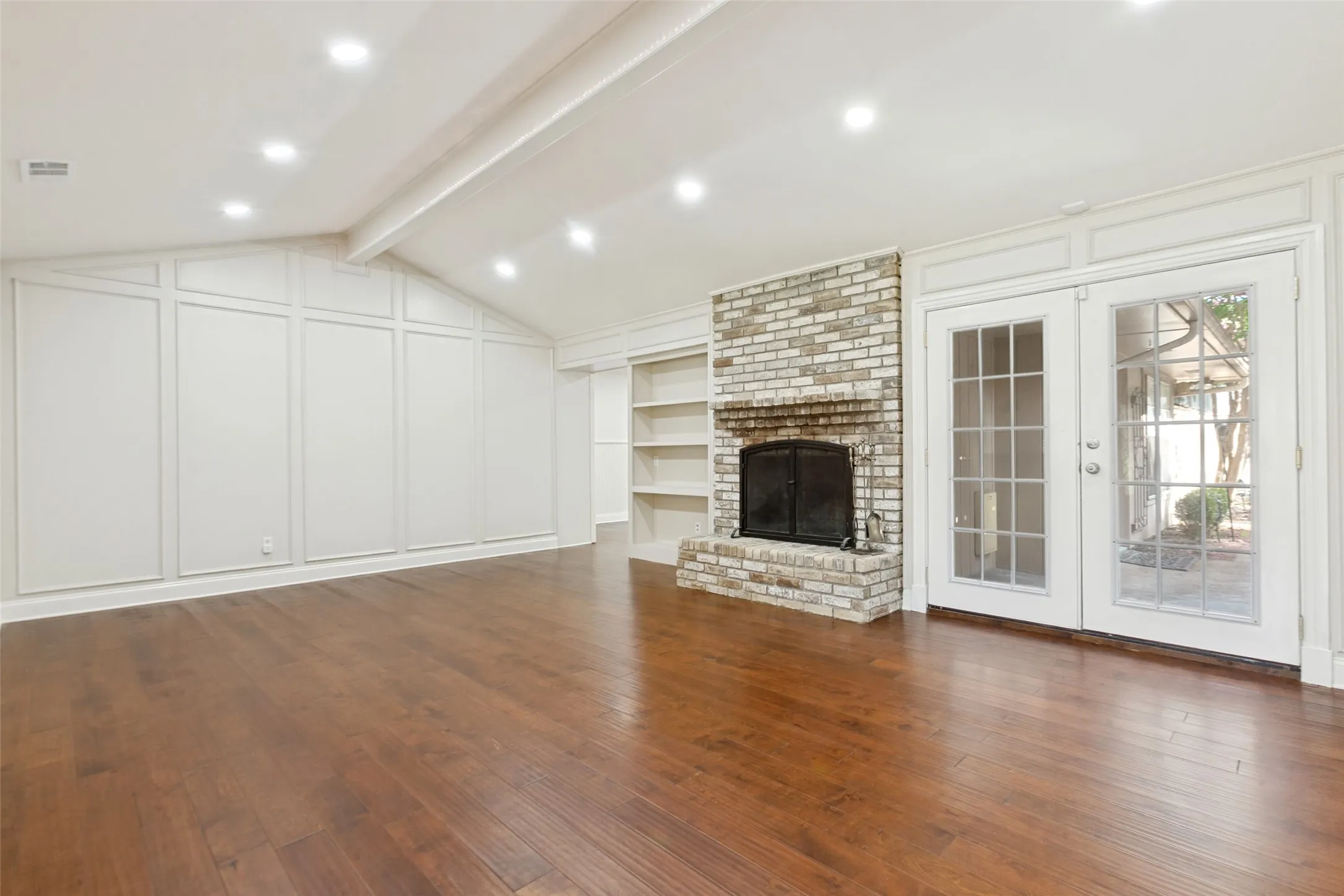 Spacious room featuring wood-finish flooring, a brick-surround fireplace, built-in shelving, vaulted ceilings with recessed lighting, and French doors