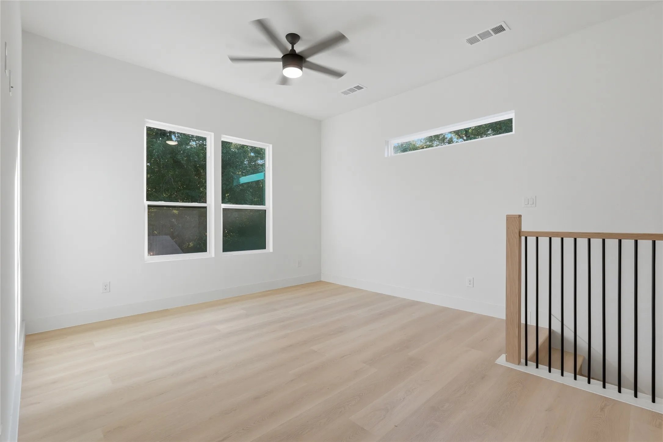 Spacious room featuring light wood-finish flooring, a contemporary ceiling fan with integrated lighting, and multiple windows including a high horizontal window for natural light