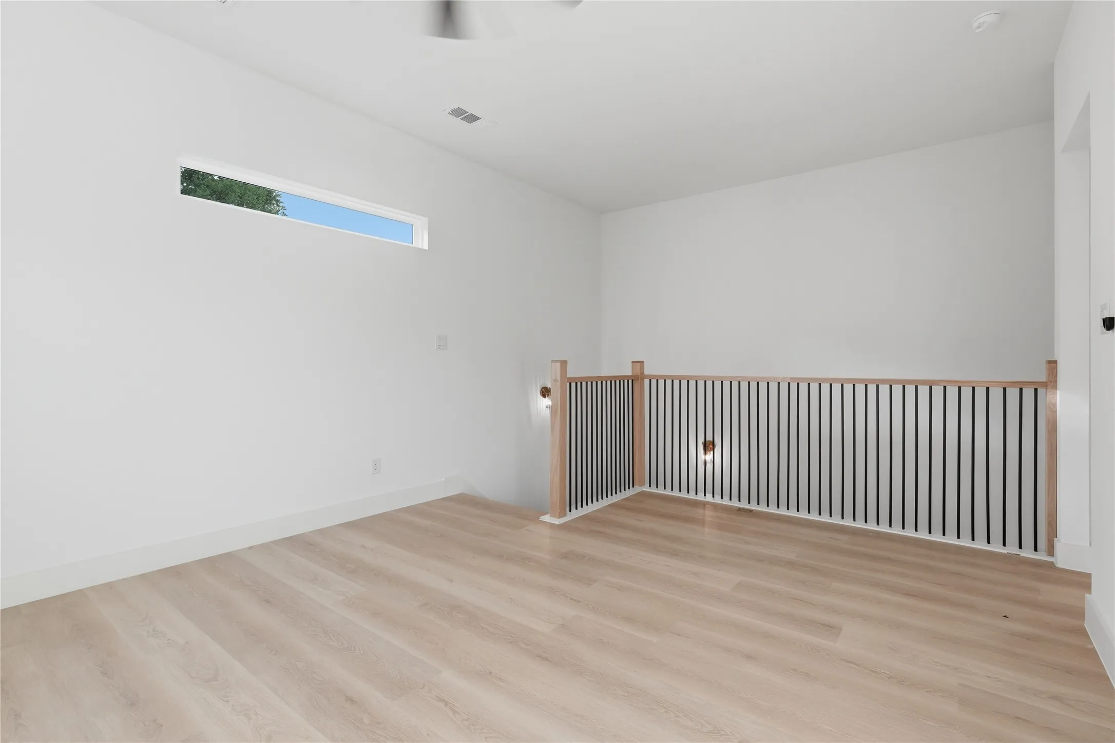 Open upper-level space featuring light wood-finish flooring, a contemporary railing with vertical black balusters and wood handrails, and a high horizontal window