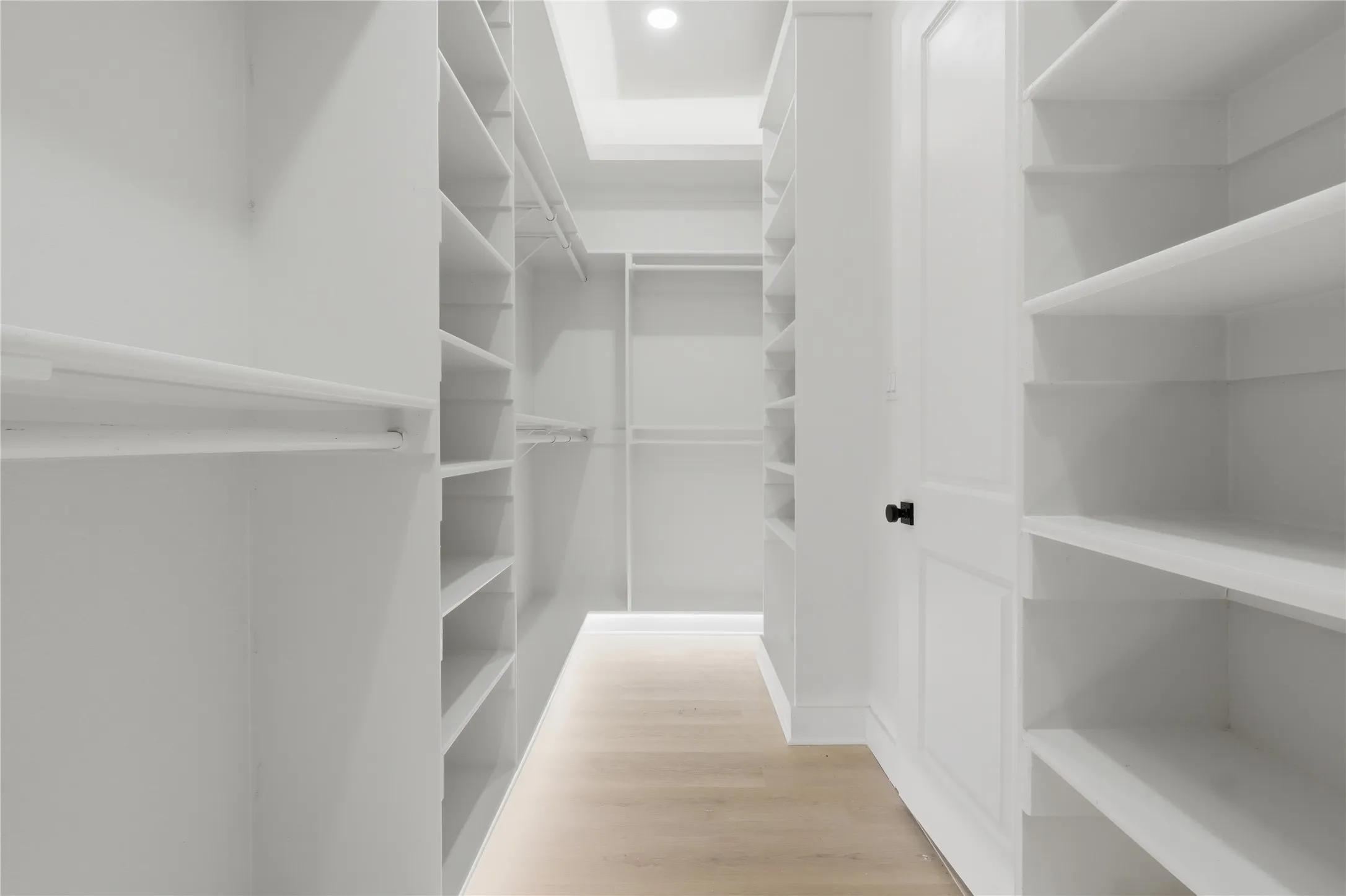 Spacious walk-in closet featuring extensive built-in shelving, multiple hanging rods, clean white finishes, light wood-finish flooring, and recessed ceiling lighting