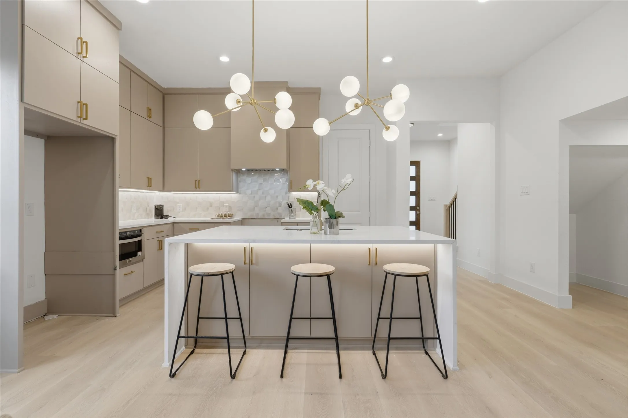 Spacious kitchen featuring a large central island with integrated lighting, two contemporary globe chandeliers, light-colored wood-finish flooring, ample cabinetry with gold-tone hardware, and a tiled backsplash