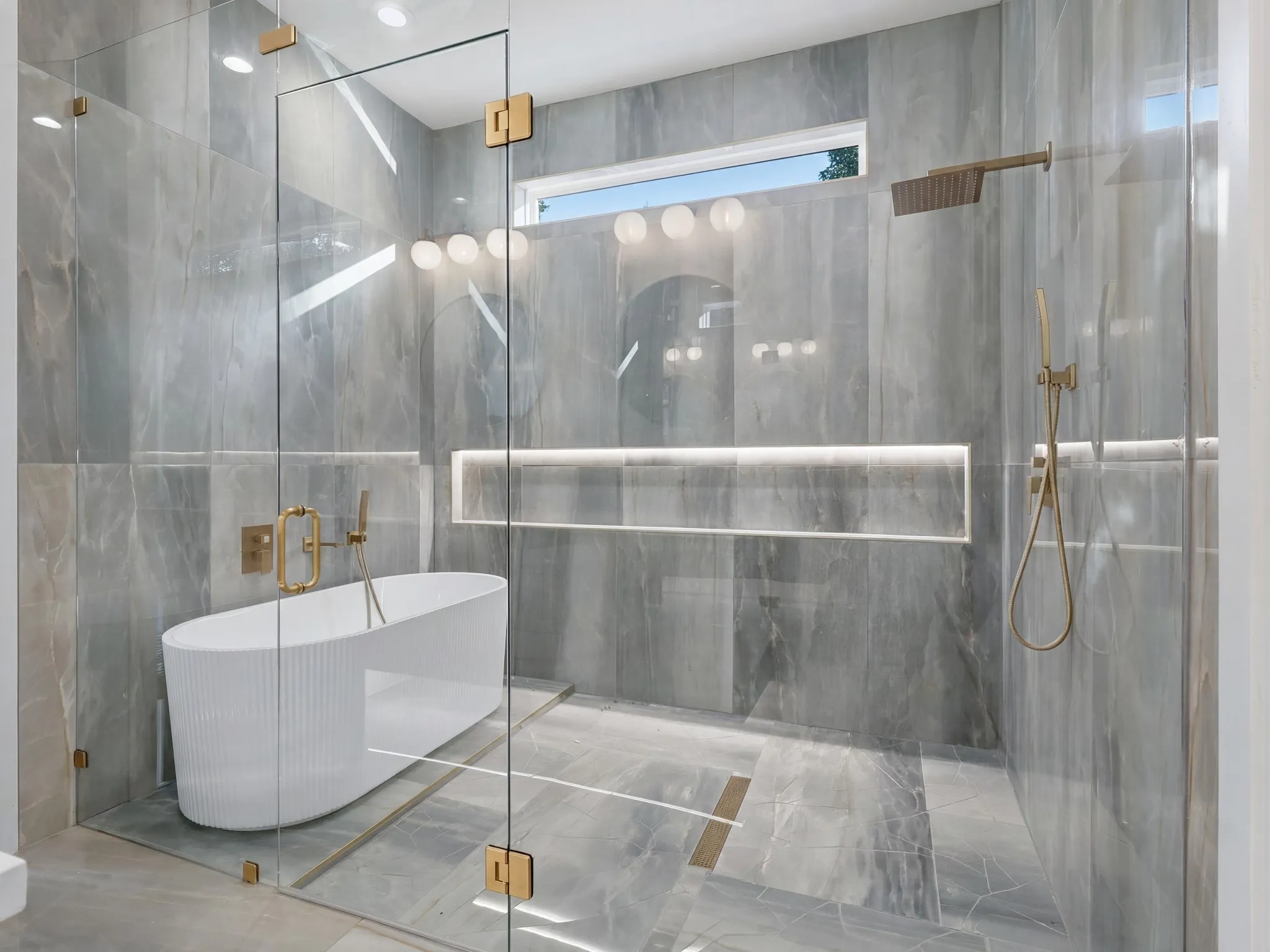 Spa-inspired bathroom featuring a freestanding tub within a glass-enclosed wet room, large format grey tile flooring and wall surround, a wall-mounted showerhead, and recessed shelf lighting