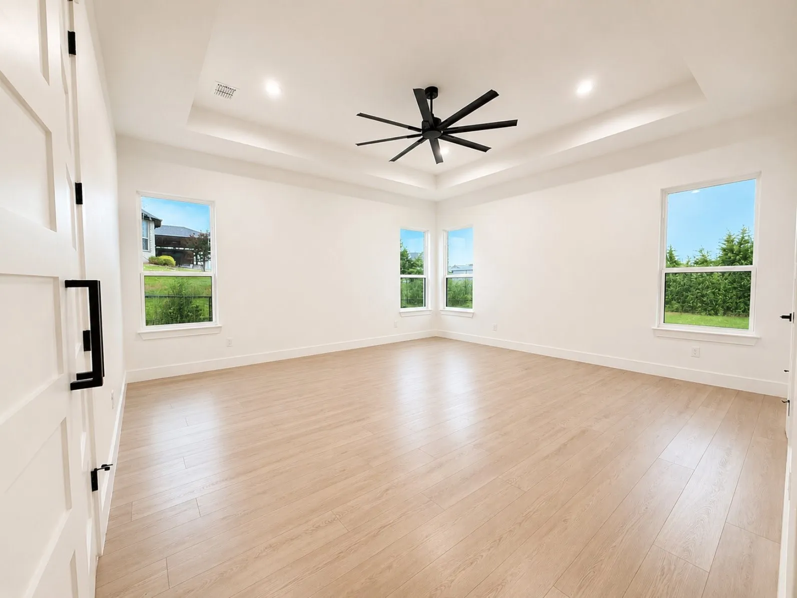 Spacious room featuring light wood-finish flooring, a tray ceiling with recessed lighting, a modern black ceiling fan, and multiple windows
