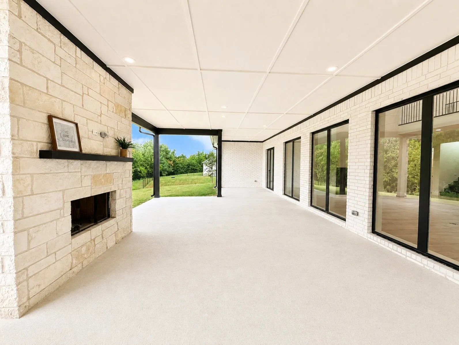 Expansive covered patio featuring a stone fireplace with a dark wood mantel, recessed lighting, white brick exterior walls, and multiple large black-framed glass windows