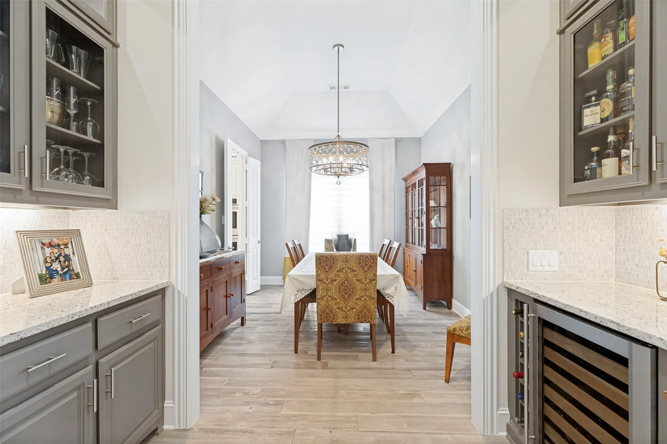 Special family occasions and gracious entertaining will be enjoyable in the elegant formal dining room.  It is accessed through a through a short hallway equipped with  a wine bar with cabinets on one side and a wine fridge on the other.  Granite countertops are topped with stacked stone backsplashes.  The LVP flooring flows into the dining room where a stunning chandelier glows from the raised ceiling.  Tall windows have shades and are dressed with pretty drapery panels.