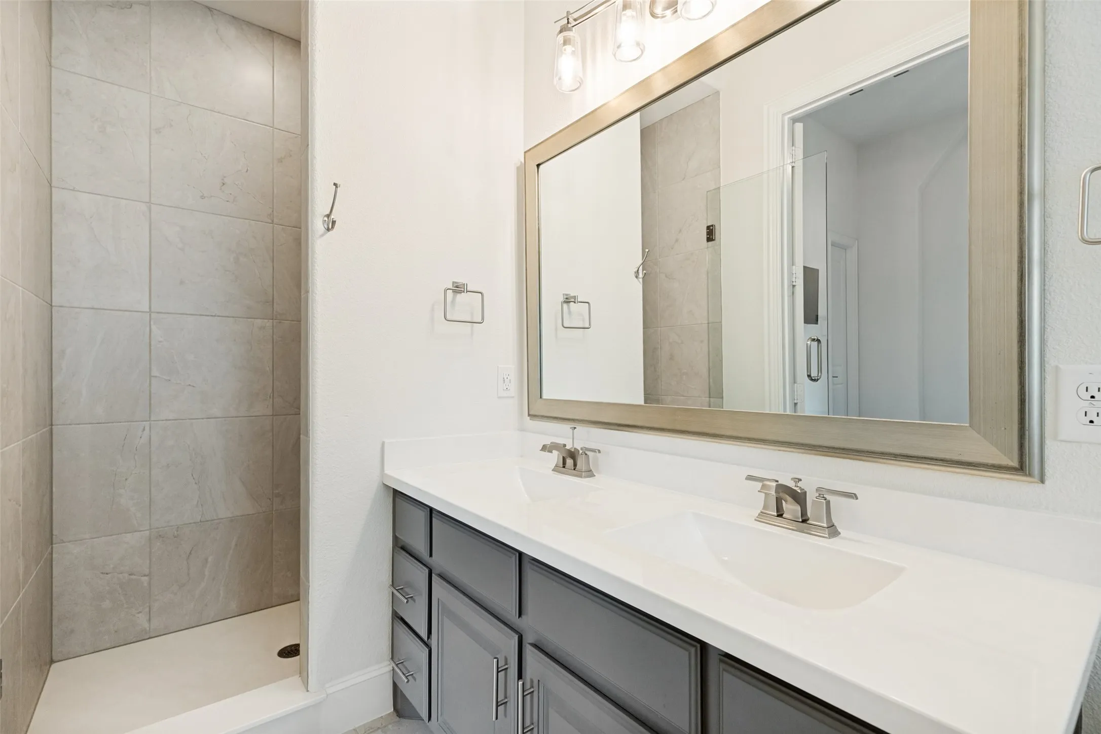 This bathroom is en-suite for the second downstairs bedroom.  With guests in mind, the bath was thoughtfully designed with a vanity with two integrated sinks in the cultural marble countertops.  A wide metallic framed mirror is above and light fixtures above the mirror add light.  A roomy shower is enclosed with floor tile, and ceramit tile is on the floor.