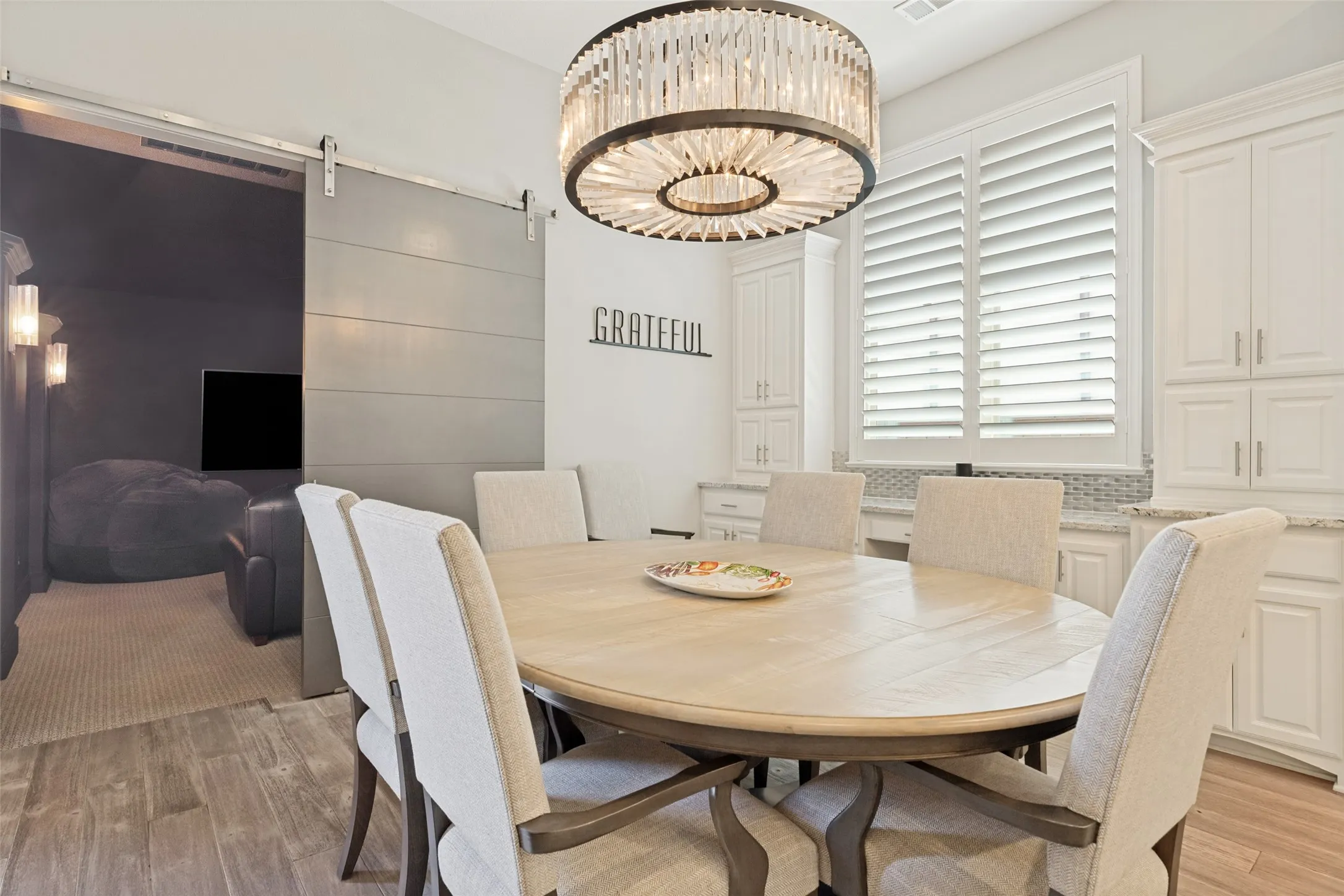 This casual dining area is ideal for family meals and casual entertaining.  The cabinets from the kitchen extend into the dining area and include a desk which is handy for homework or daily planning.  A window with shutters is above.  A striking chandelier adds an elegant touch, and the Luxury Vinyl Plank floors area in this area.  Just beyond is the media room accessed by a barn door.